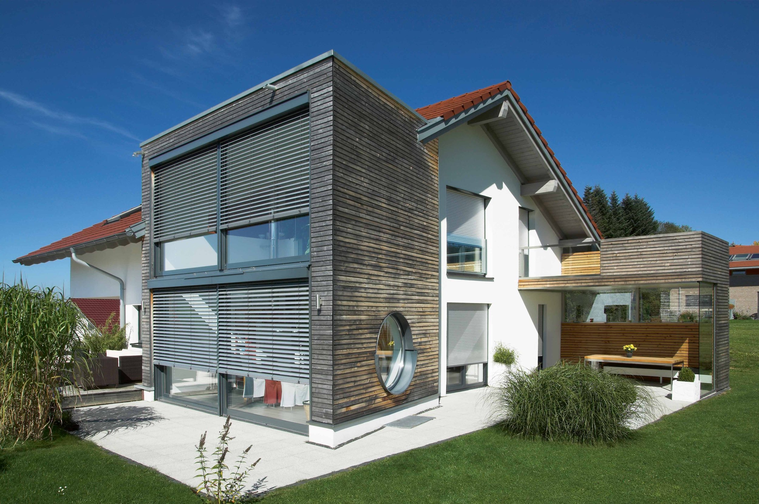Modernes zweistöckiges Haus mit Holz und weißen Wänden, großen Fenstern mit Rollos, einer Terrasse mit Gartenmöbeln und einem grünen Garten bei sonnigem Himmel.