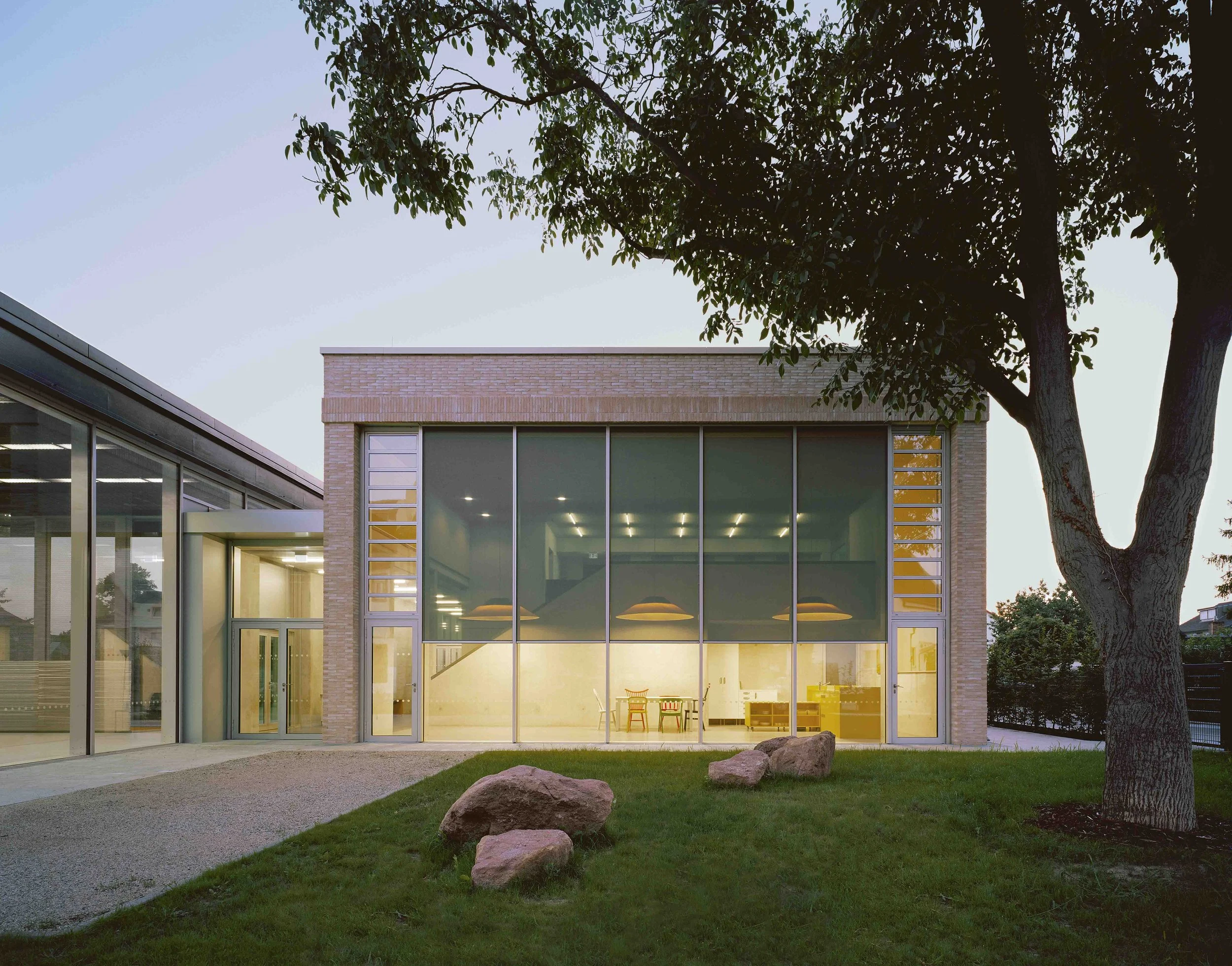 Moderne Schule mit großen Fenstern, umgeben von Rasen, Steinen und einem Baum bei Dämmerung.