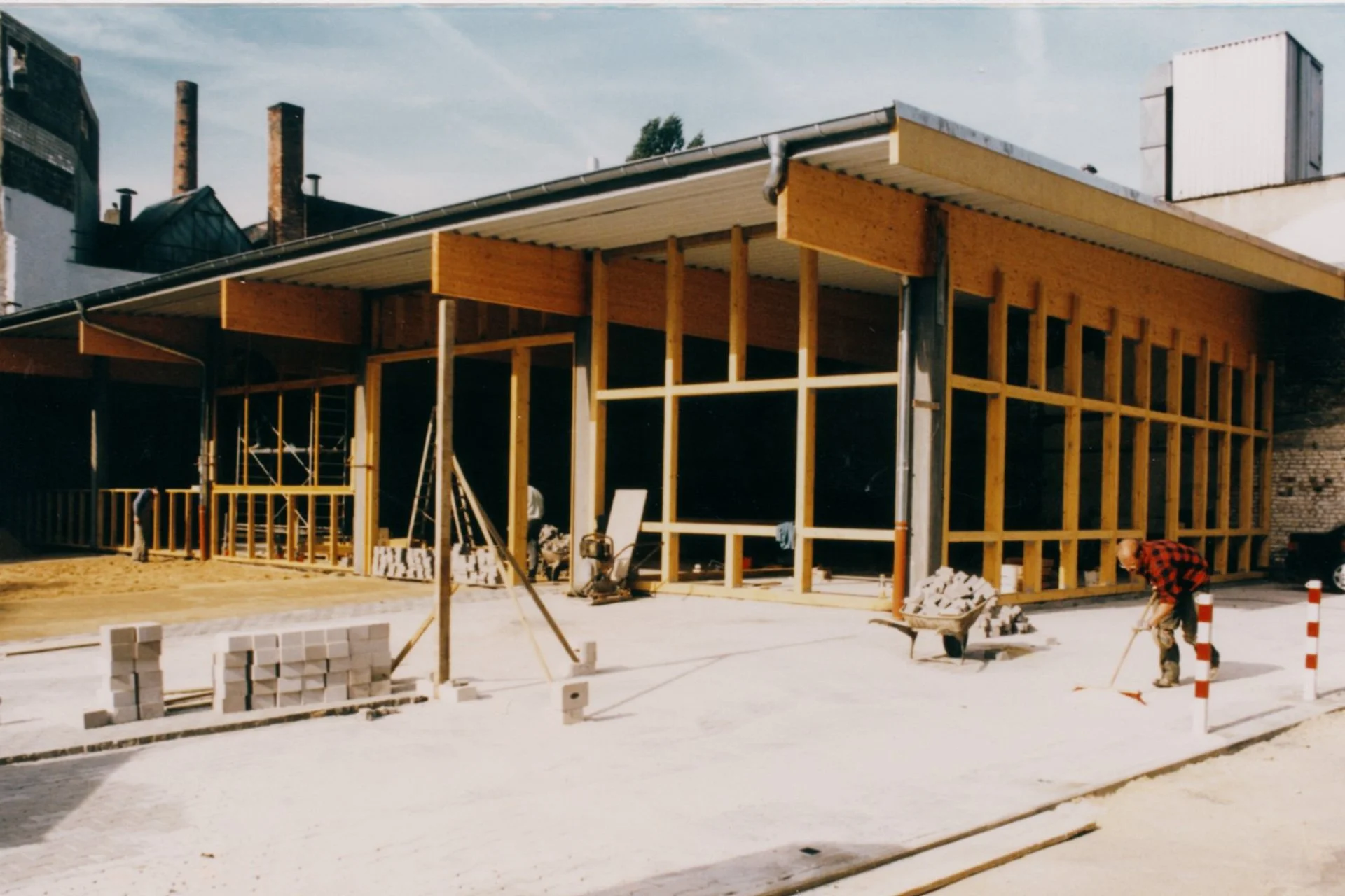 Bauarbeiter bauen an einem Gebäude mit Holzrahmen auf einer Baustelle, Steine und Werkzeuge sind sichtbar.