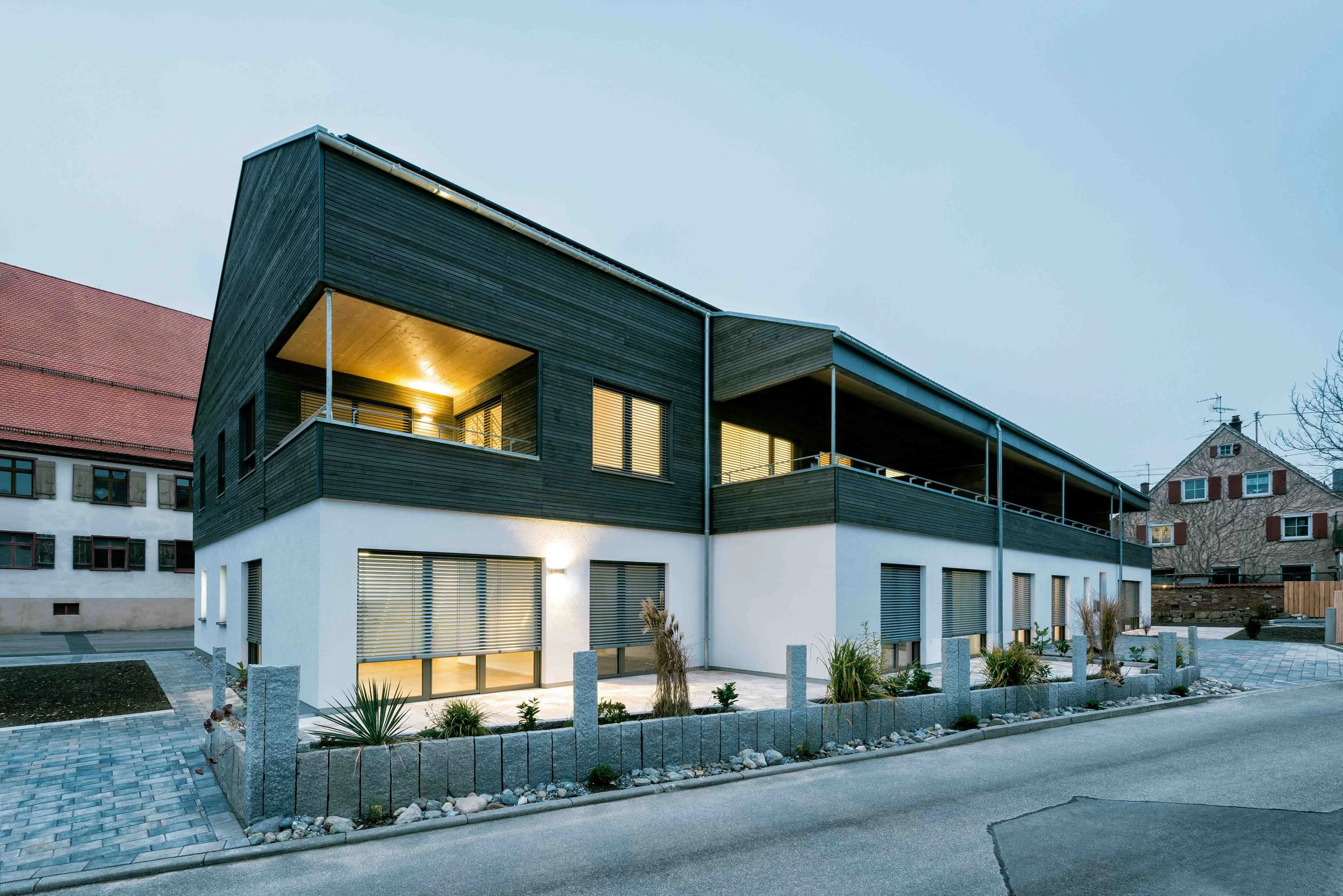 Modernes zweistöckiges Haus mit weißen und schwarzen Elementen, große Fenster, Balkon und eingeschaltete Innenbeleuchtung, umgeben von einem steinernen Zaun und minimal gestaltetem Garten.