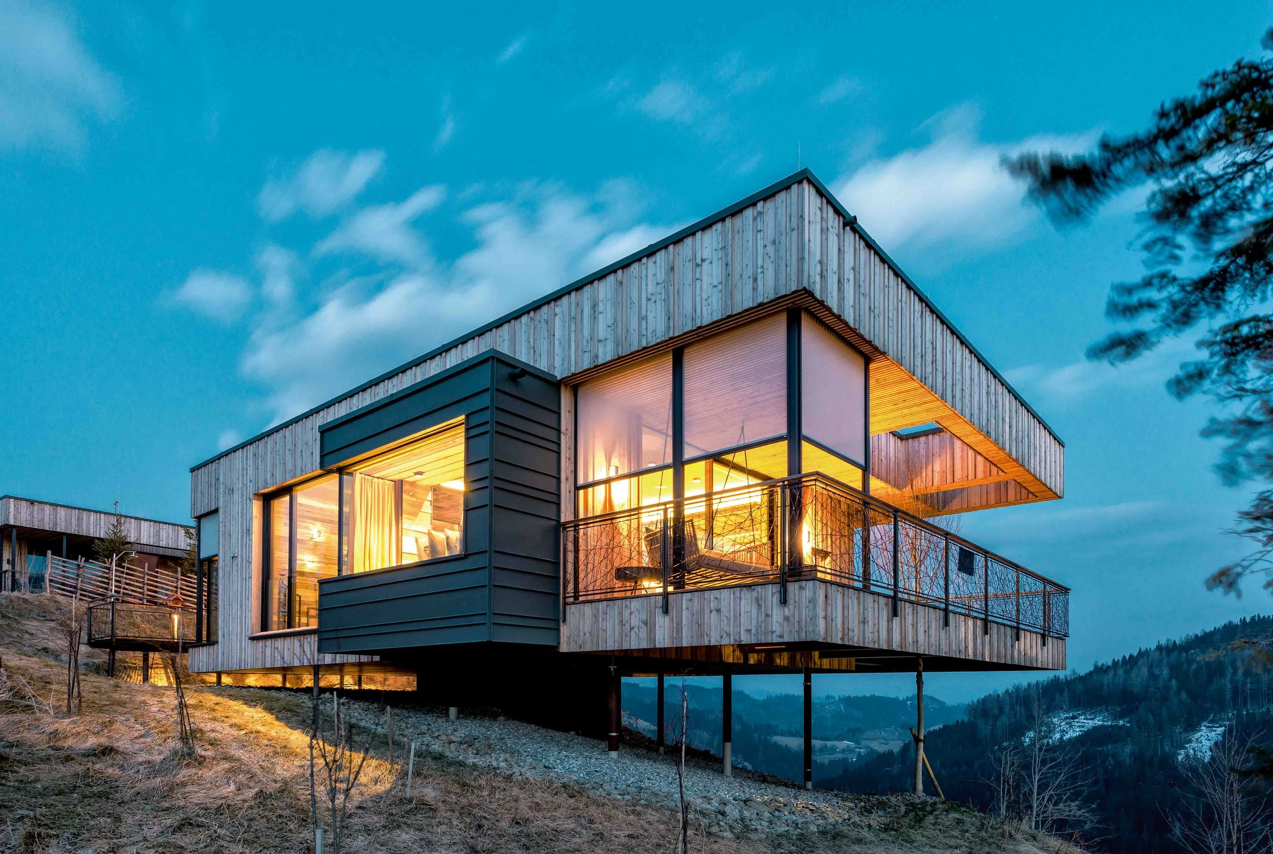 Ein modernes Haus auf einer Berghöhe bei Dämmerung mit beleuchteten Innenräumen, umgeben von Natur und Bäumen.