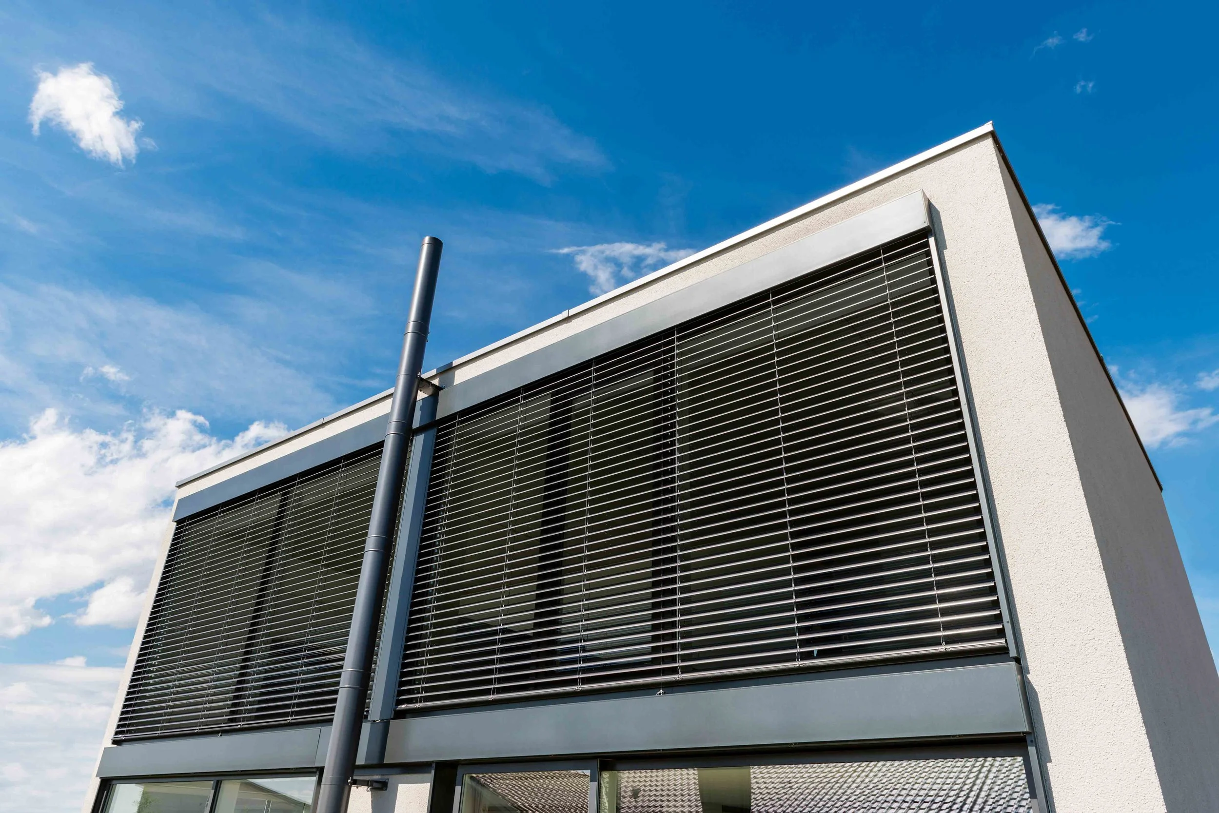 Moderner Bau mit großen Fensterfronten und Rollläden, blauer Himmel mit wenigen Wolken