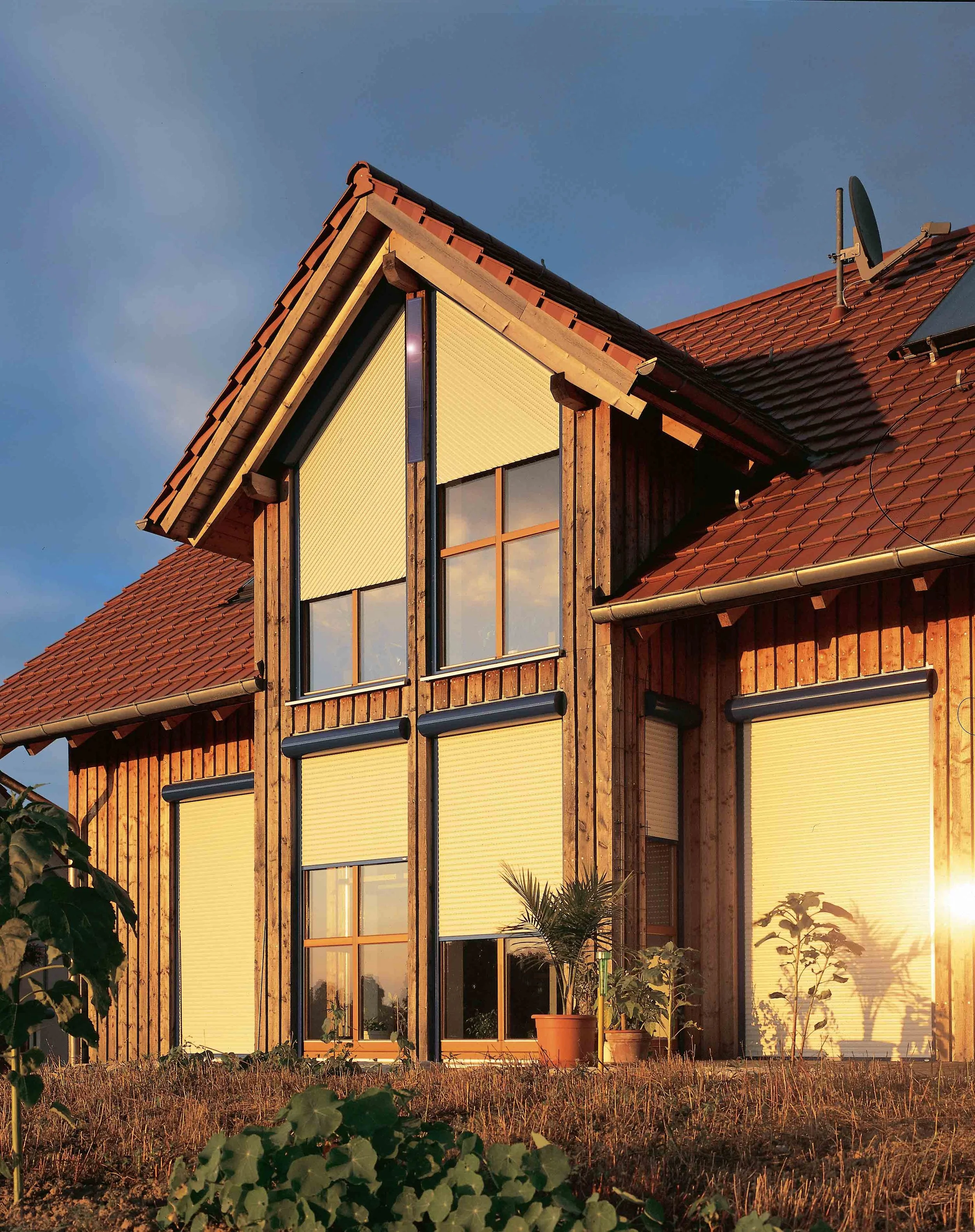 Ein modernes Holzhaus mit großen Fenstern, teilweise mit Rolläden, bei Sonnenuntergang.
