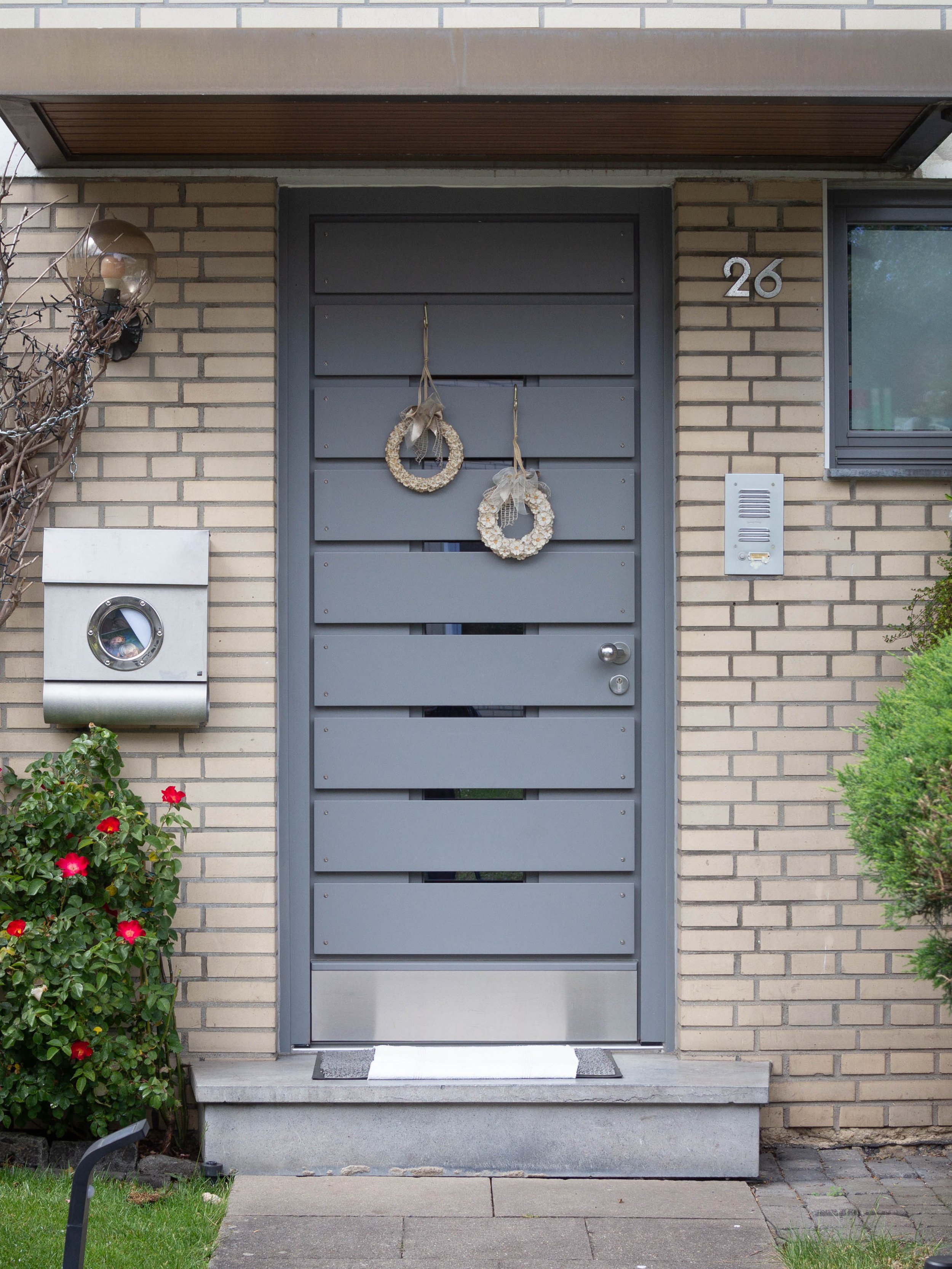 Eingangstür eines Hauses mit Hausnummer 26, geschmückte Tür mit zwei Dekorationsringen, seitliche Briefkästen, Pflanzen und Beleuchtung