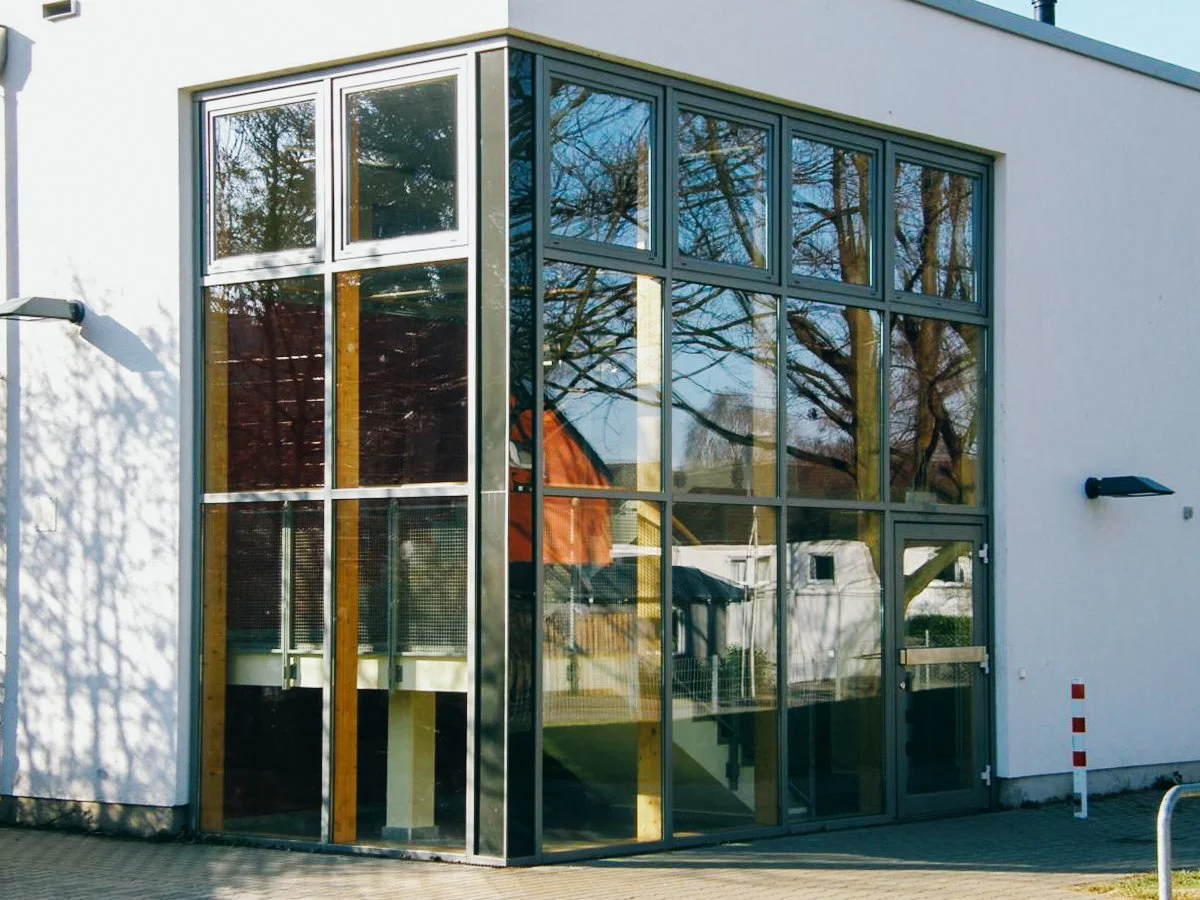 Modernes Gebäude mit großen Glasscheiben und Metallrahmen, reflektierende Fenster, umgeben von Bäumen.
