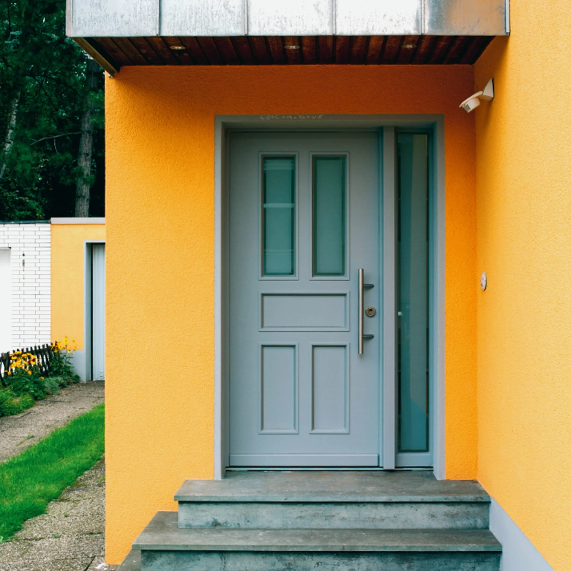 Ein blauer Eingangstür mit drei kleinen Fenstern, umgeben von gelber Hauswand, mit Steinstufen davor und grünem Garten auf der linken Seite.