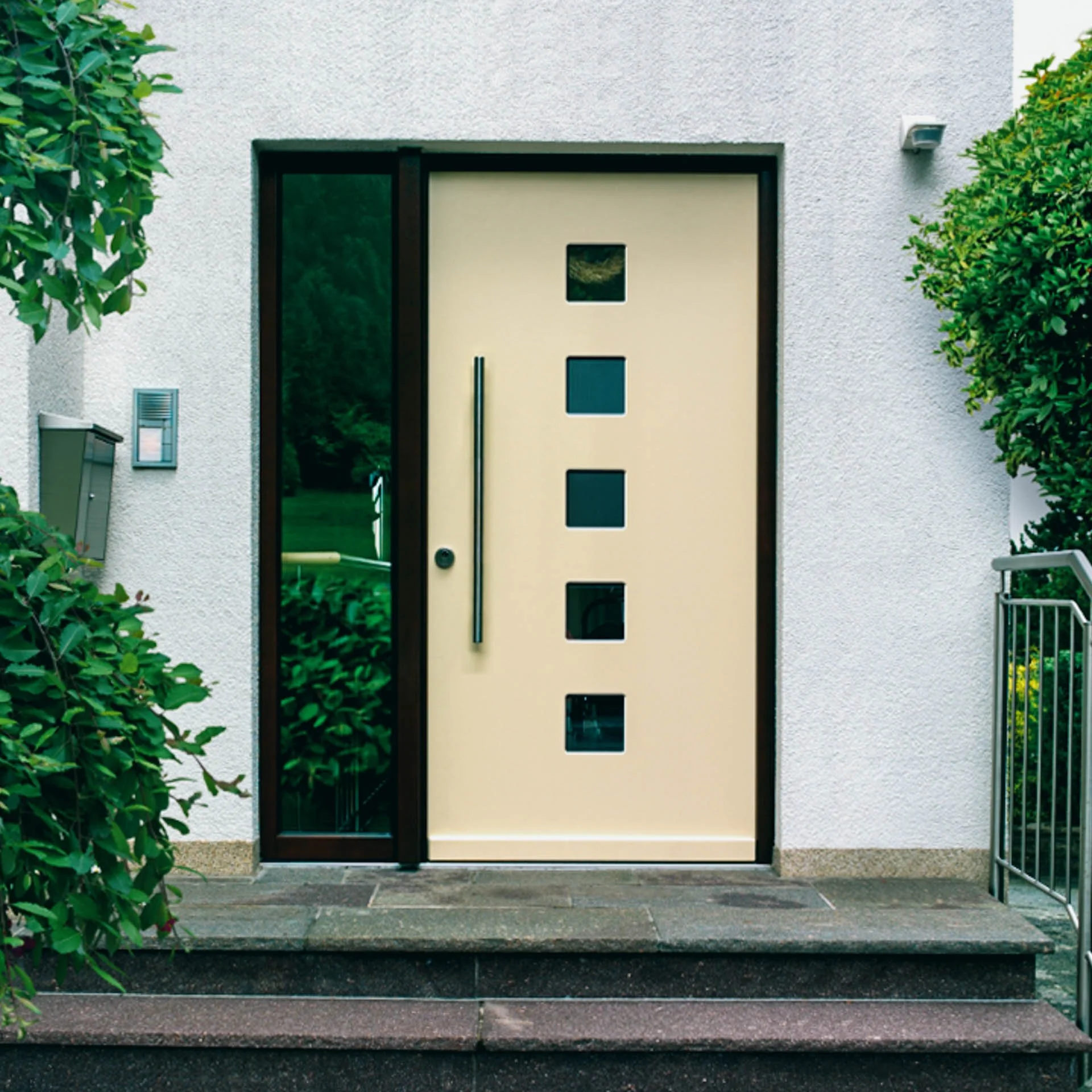 Moderner Hauseingang mit einer cremefarbenen Tür, vier kleine quadratische Fenster und seitlichem Glaselement, umgeben von grünen Büschen