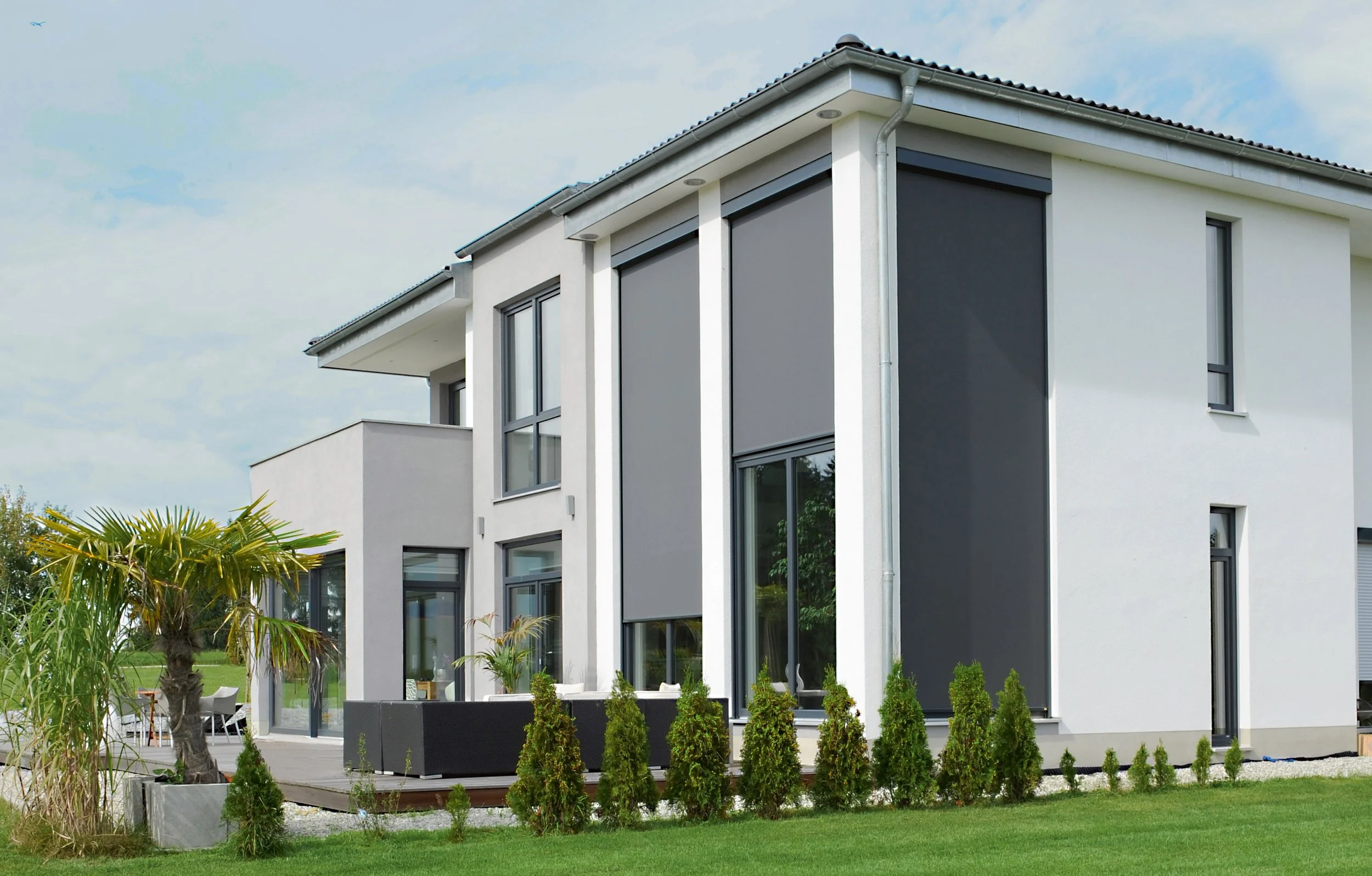 Modernes, zweistöckiges Haus mit weißen Wänden, großen Fenstern und grauen Rollläden, umgeben von grünem Rasen und kleinen Bäumen.