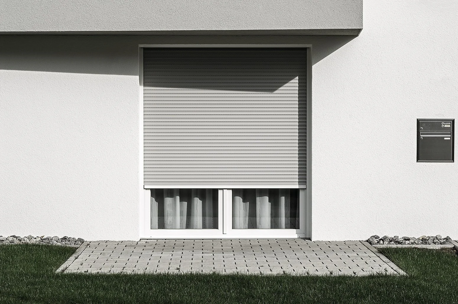 Weiße Hausfassade mit grauem Rollladen vor Fenster, daneben Briefkasten auf der rechten Seite, vor dem Haus ein gepflasterter Eingangsbereich, umgeben von grünem Rasen.