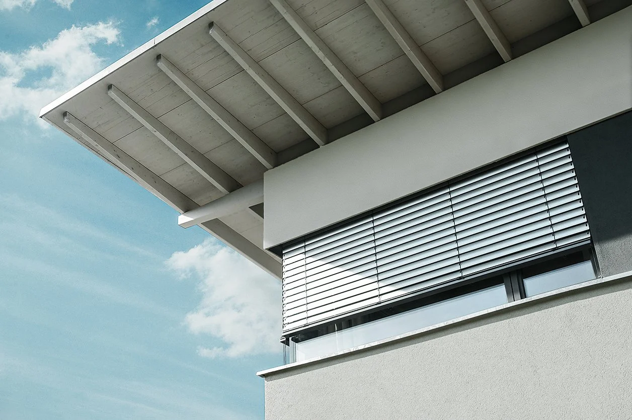 Ein modernes Haus mit einer schattigen Terrasse und Sonnenblenden vor einem blauen Himmel mit weißen Wolken.