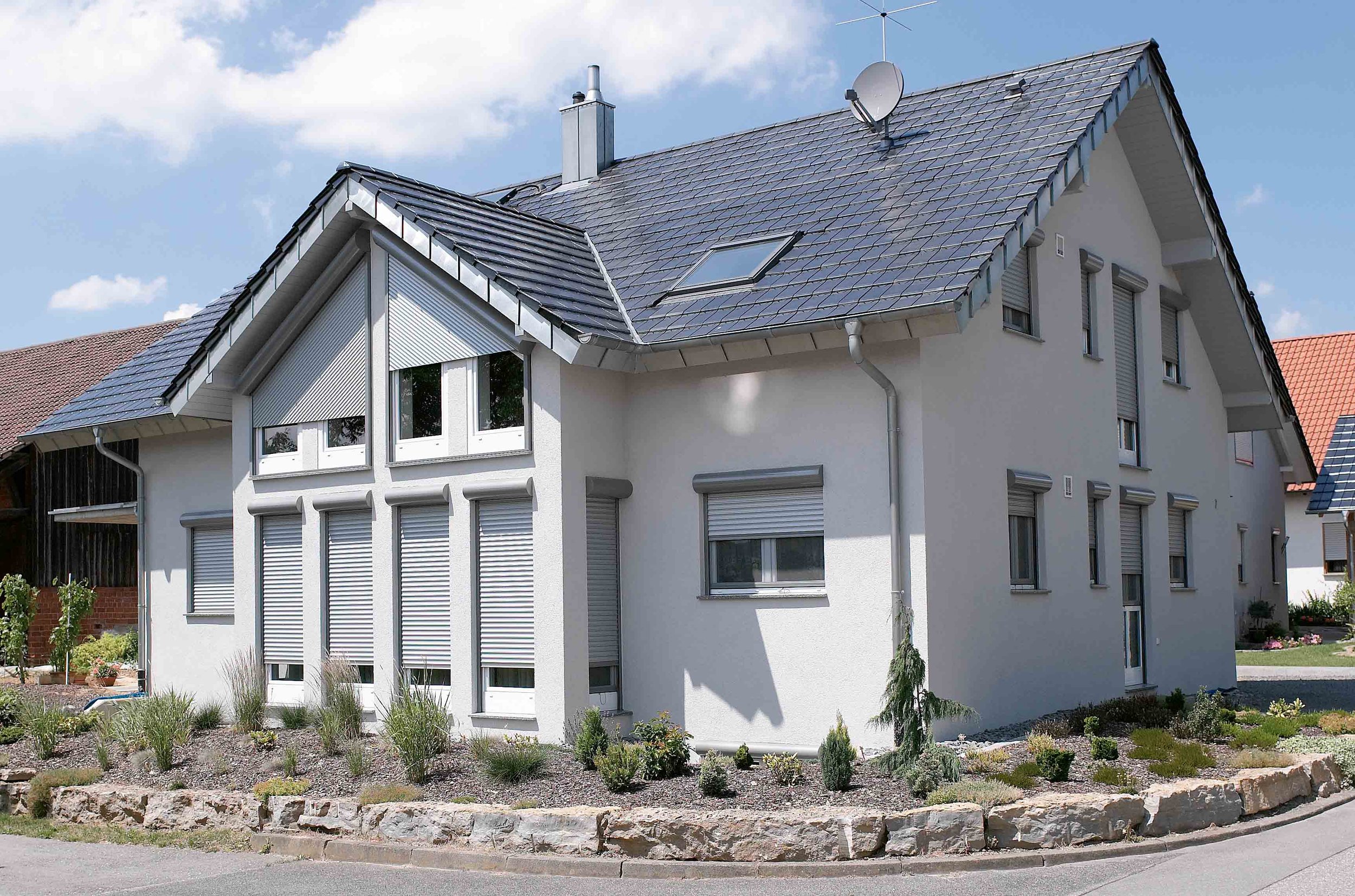 Ein modernes, weiß gestrichenes Mehrfamilienhaus mit Satteldach, Dachfenstern und Rollläden, umgeben von einem kleinen Garten mit Bepflanzung.