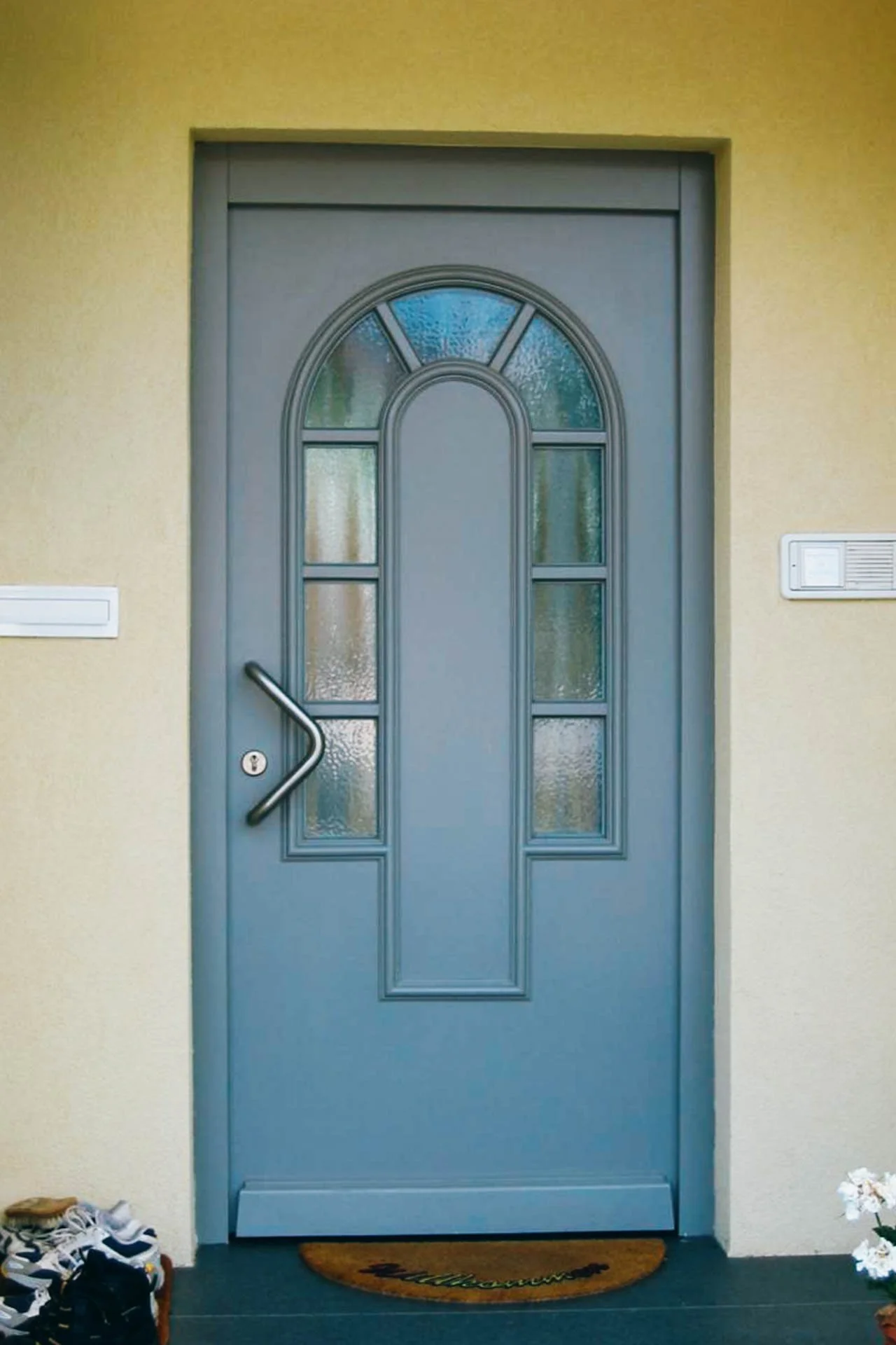 Blaue Haustür mit einem gewölbten Fenster und einem gebogenen Türgriff, vor einem hellgelben Wand