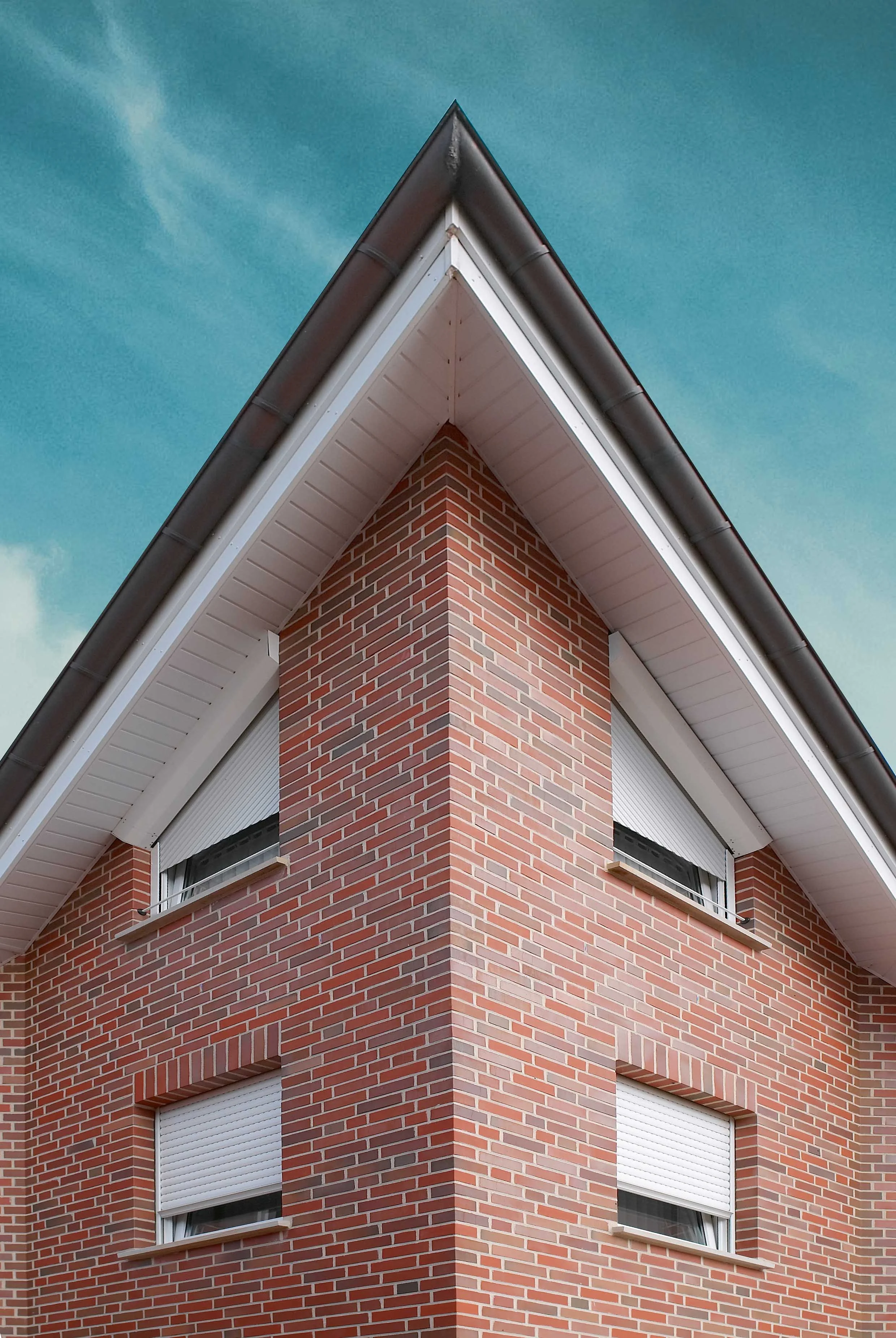 Eckhaus eines roten Backsteinhauses mit weißen Rolläden vor den Fenstern, Dach mit schwarzem Ziegel und blauem Himmel im Hintergrund.