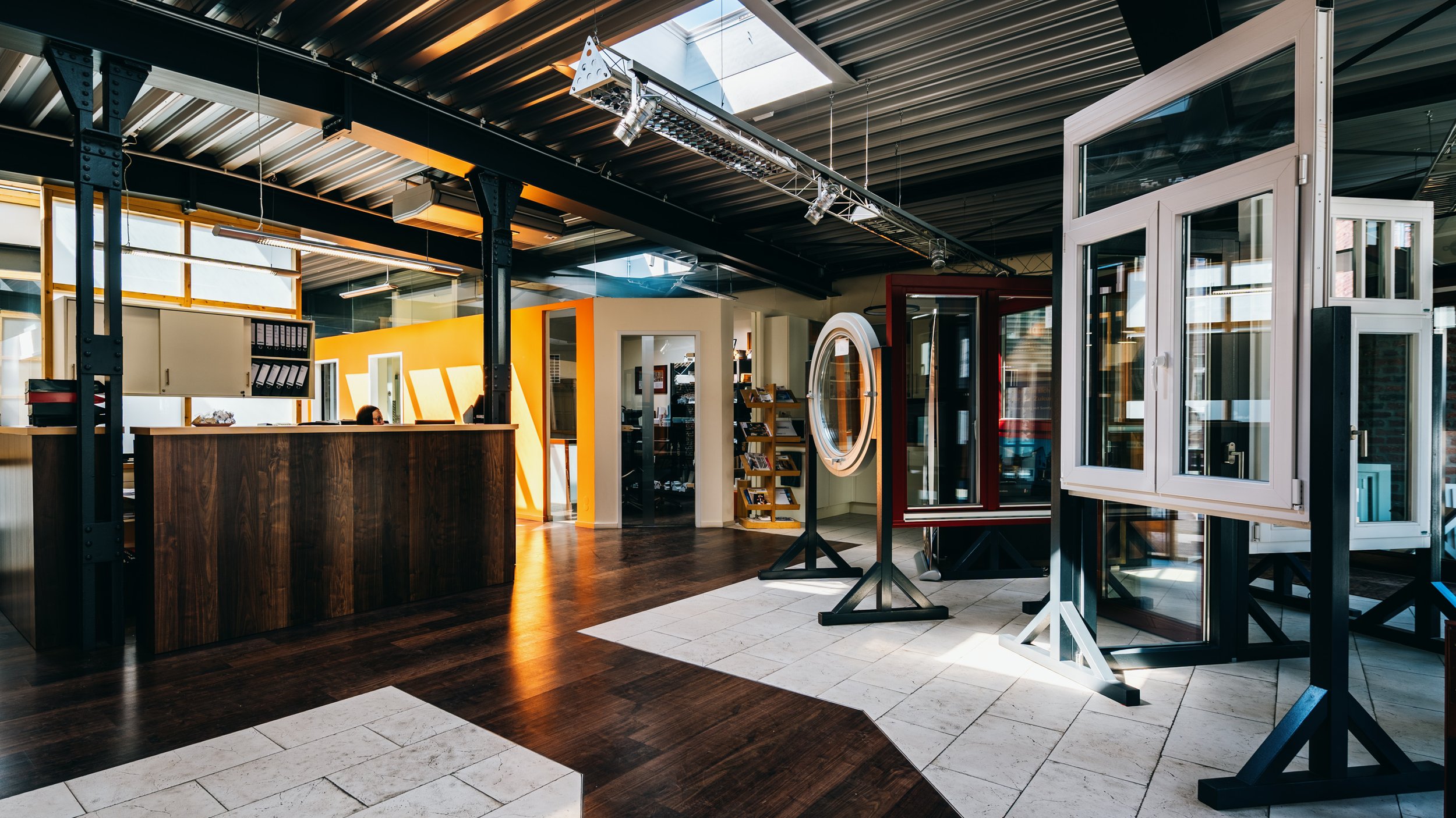 Innenansicht eines Showrooms für Fenster und Türen mit verschiedenen Modellen auf Stellständern, dunklem Holzboden und einer Empfangstheke.