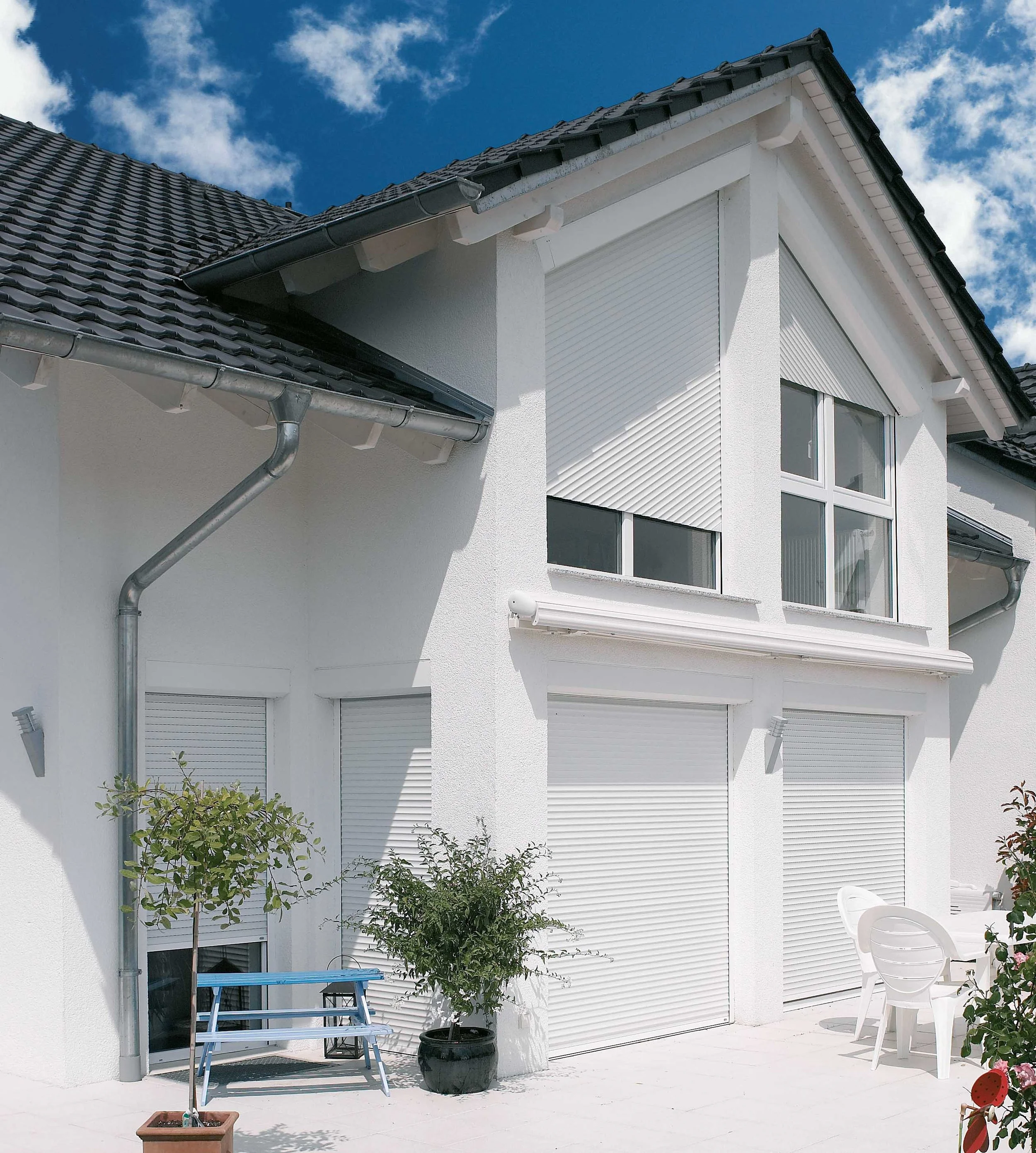 Ein modernes weißes Haus mit rollläden vor Fenstern, einem Balkon, Gartenmöbeln und Pflanzen, unter blauem Himmel.