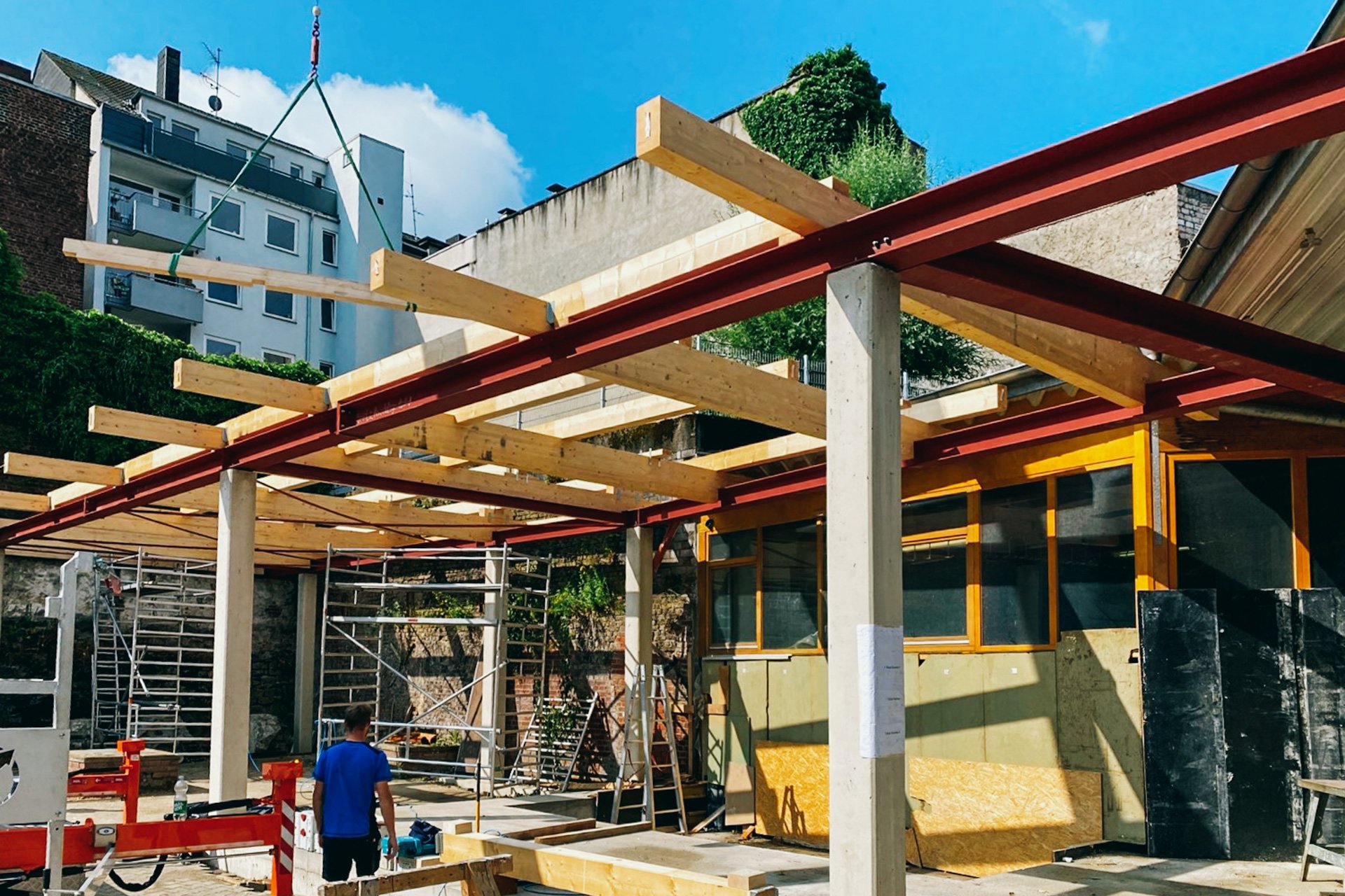 Bauarbeiten an einer Gebäudestruktur mit Holz- und Stahlträgern, Arbeiter und Baugerüst sichtbar.