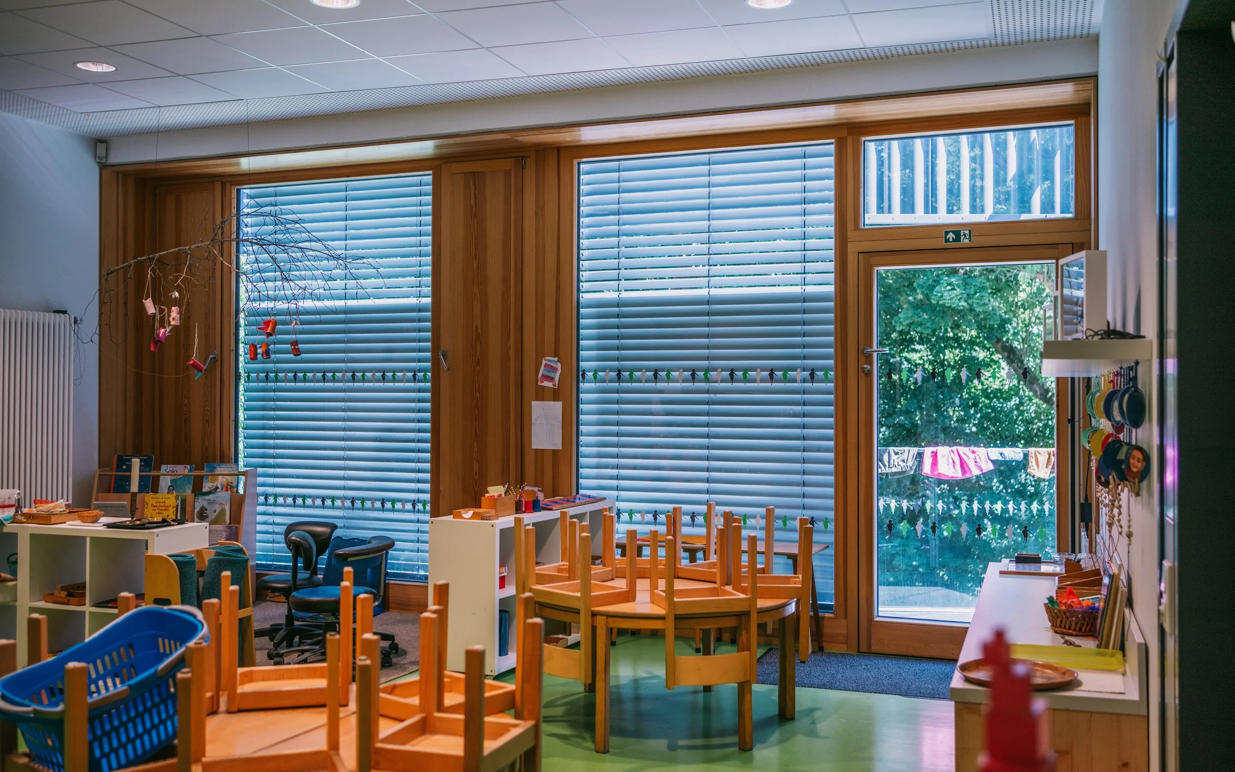 Ein ingerichtes Klassenzimmer mit umgestellten Tischen und Stühlen, größeren Fenster mit Jalousien und Blick nach draußen auf eine Wäscheleine mit bunten Kleidungstücken, dekoriert mit Schnappverschlüssen.