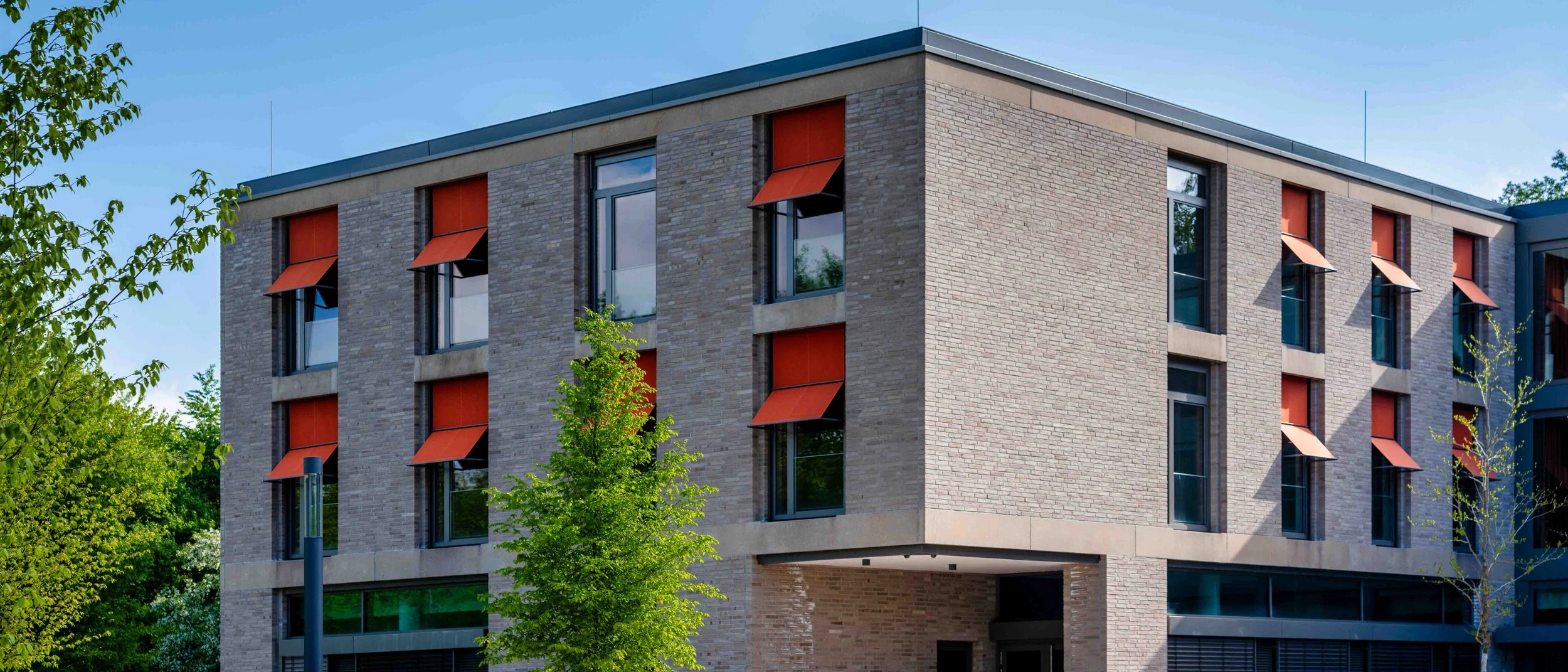 Mehrstöckiges modernes Gebäude mit großen Fenstern und roten Sonnenschutzmarkisen, umgeben von grünen Bäumen bei Tageslicht.