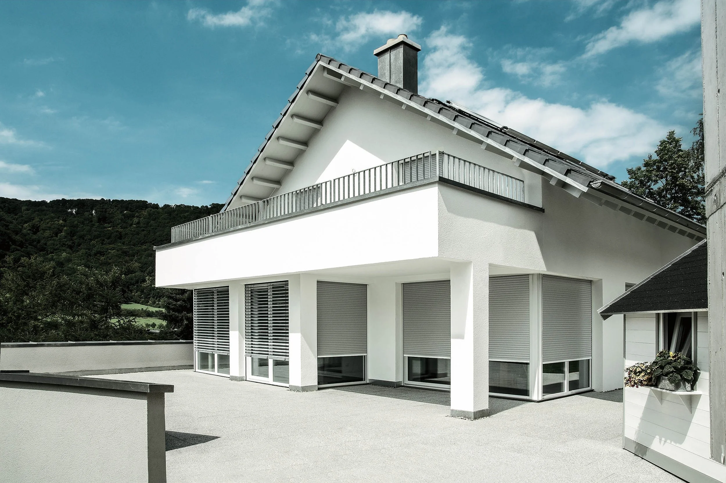 Moderner weißer Wohnhaus mit Balkon und großen Fenstern, im Hintergrund bewaldete Hügel, sonniger Tag, blauer Himmel mit Wolken.