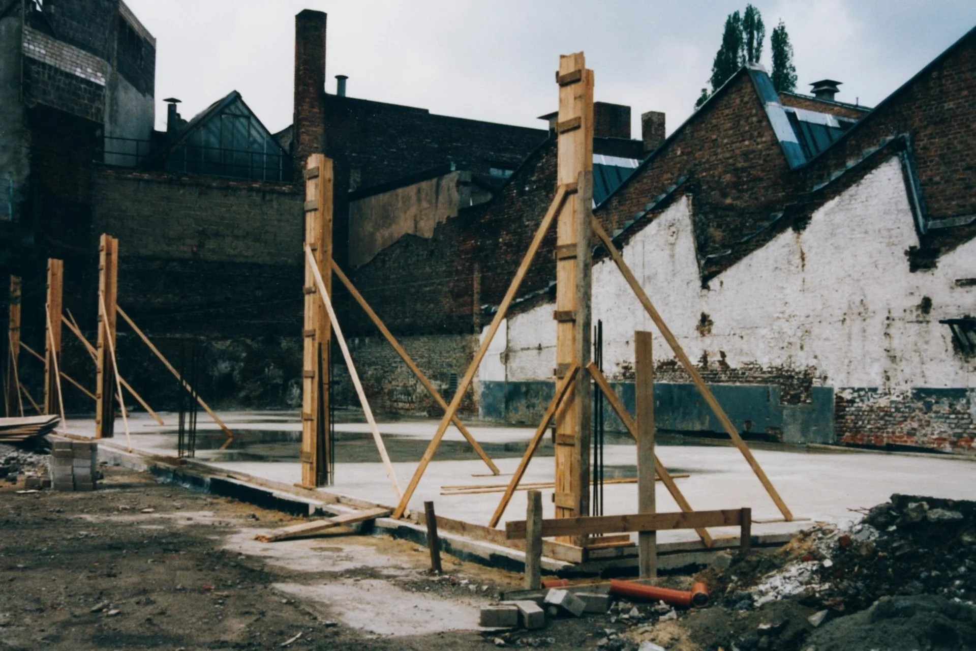 Baugrund mit Holzständer für Fundament, umgeben von alten Gebäuden mit Ziegelwänden, in einem städtischen Umfeld