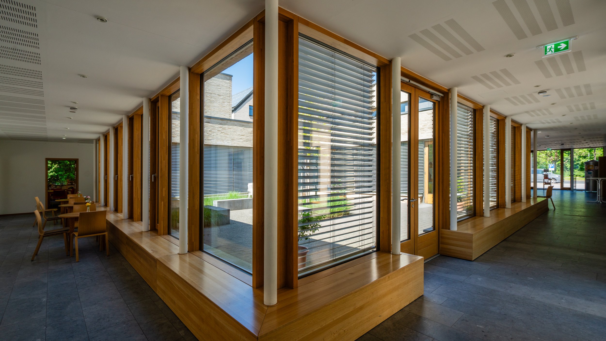 Innenraum mit großen Fenstern, Holzrahmen, Sitzbank und Tischen, beleuchtet durch Tageslicht, Flur mit Türmaterialien, Notausgangschilder.