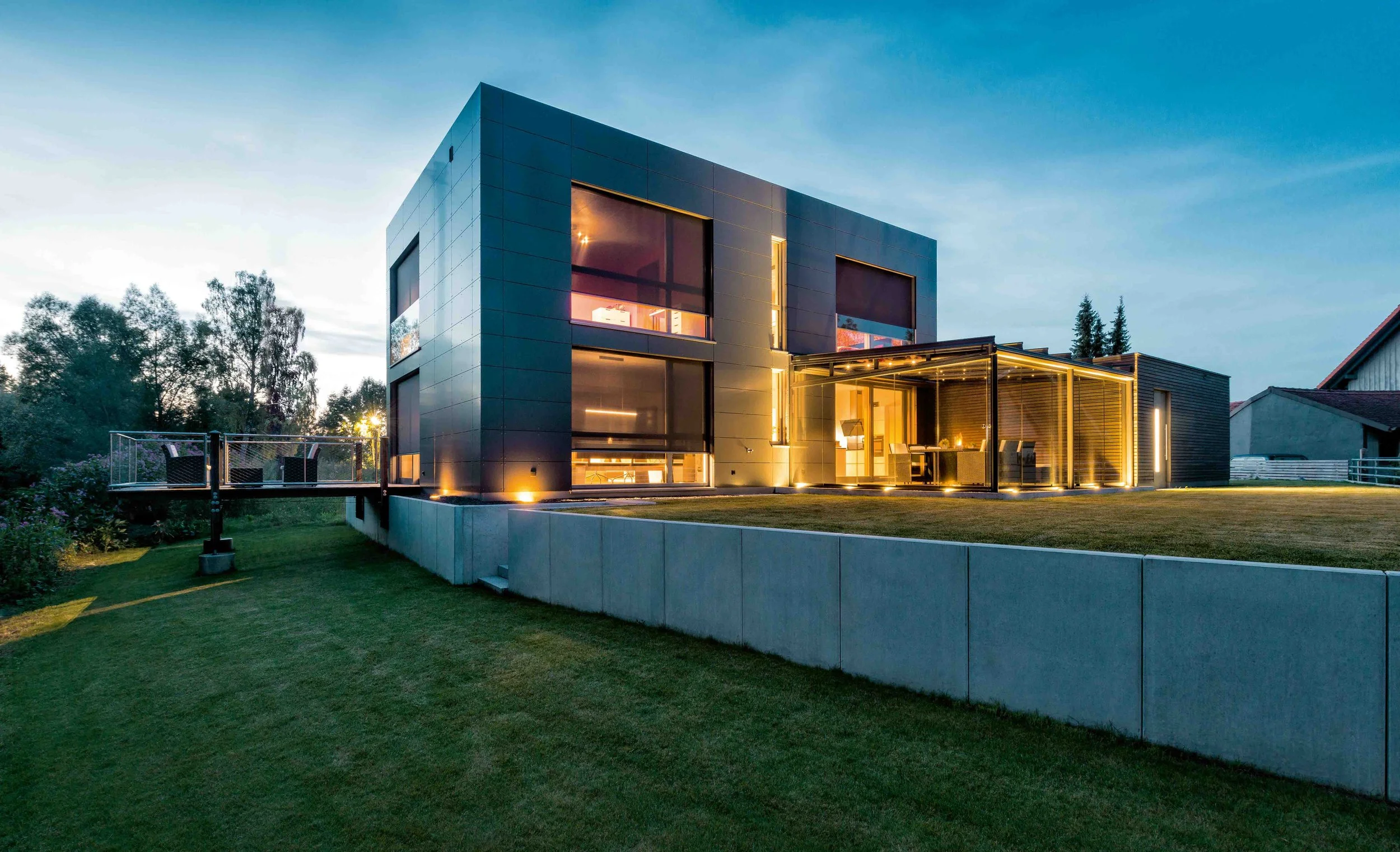 Moderne mehrstöckige Villa bei Dämmerung mit beleuchteten Innenräumen, große Fenster und gepflegte Gartenflächen.