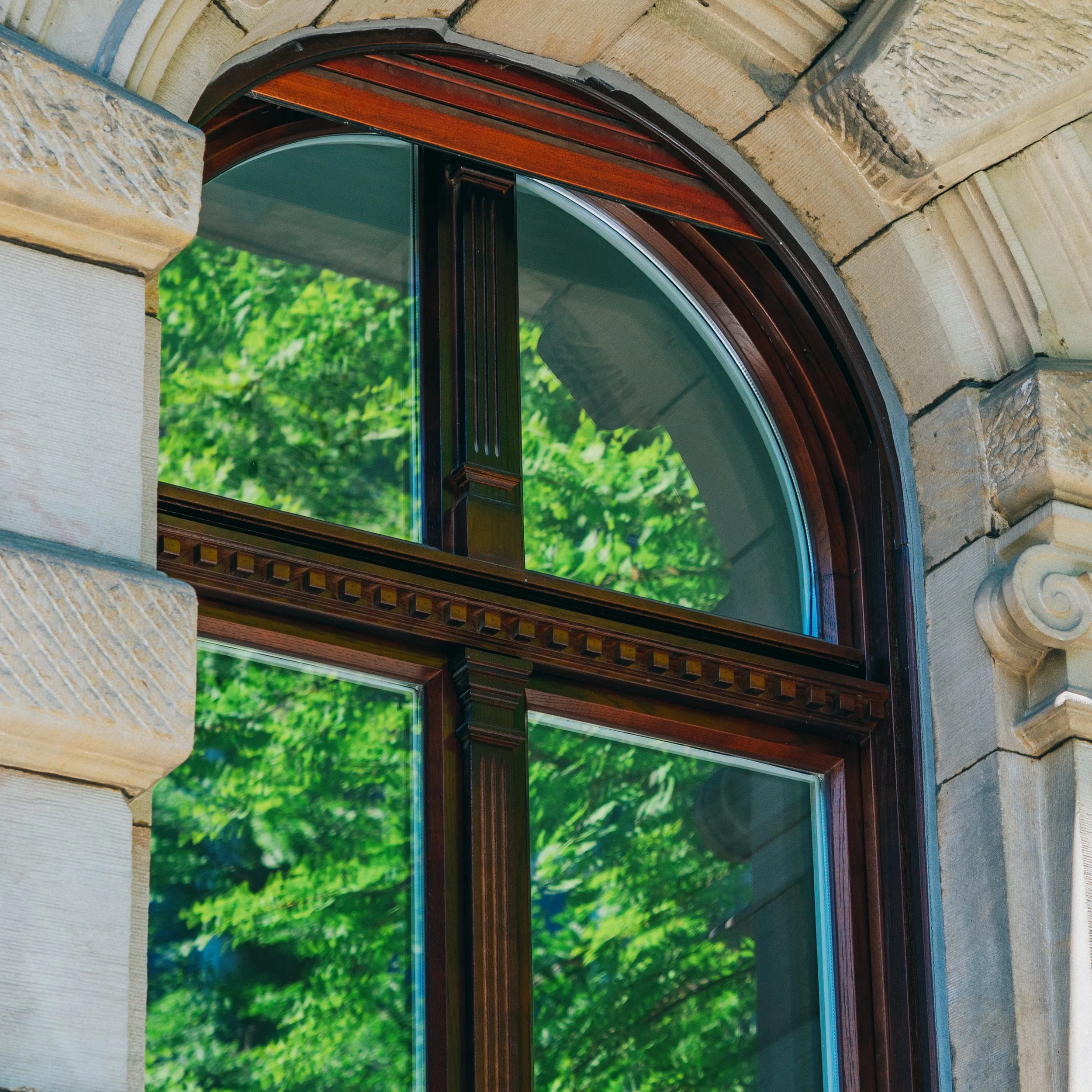 Nahaufnahme eines großen, bogenförmigen Fensters mit dunklem Holzrahmen vor einem Steinmauer-Hintergrund, mit grünen Blättern im Blick durch das Fenster.