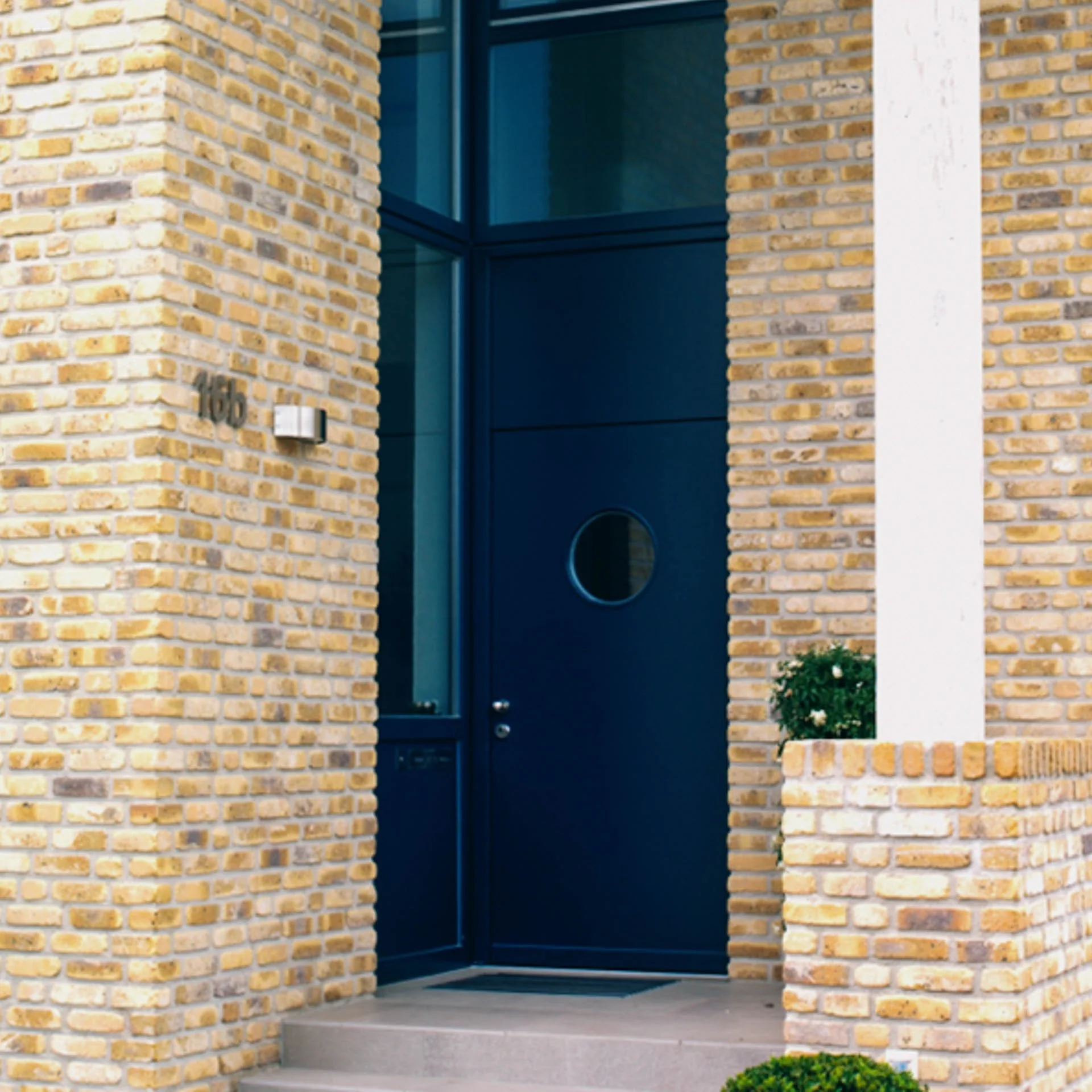 Blick auf die Eingangstür eines Gebäudes mit gelben Backsteinwänden und einer blauen Tür mit kleinem runden Fenster, umgeben von weißen Wandabschnitten und kleinen Pflanzen.