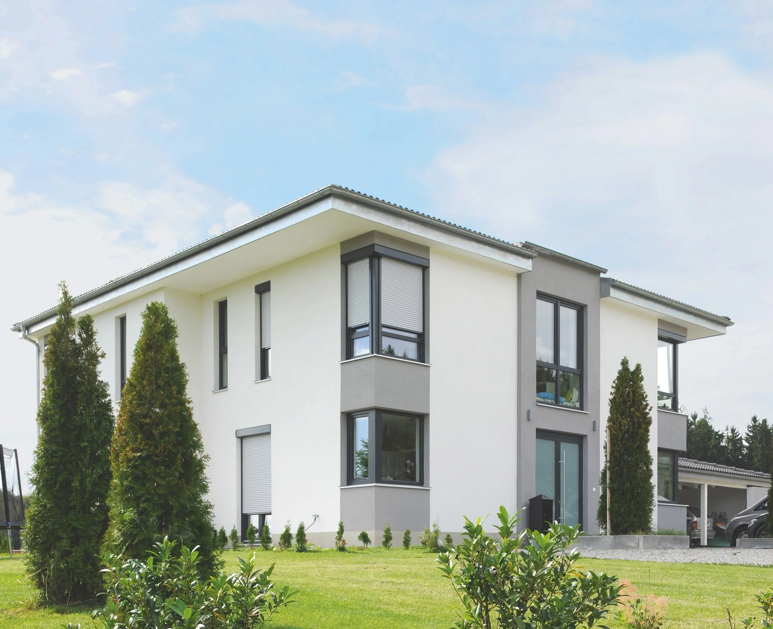 Ein modern zweistöckiges Haus mit weißen und grauen Wänden, großen Fenstern und einer gepflegten grünen Gartenzone mit Bäumen und Büsche.