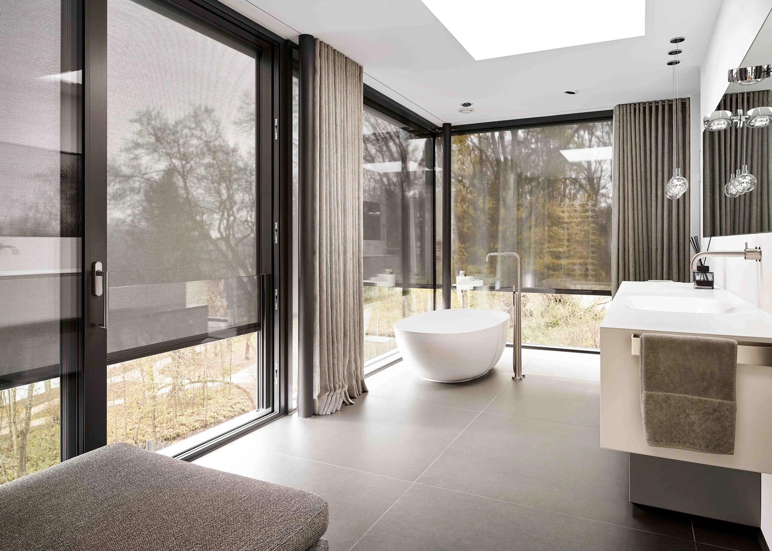 Moderne Badezimmer mit freistehender Badewanne, großen Fenstern mit Sicht auf die Natur, dunklen Vorhängen, Waschbecken mit Handtuch, und moderner Beleuchtung.