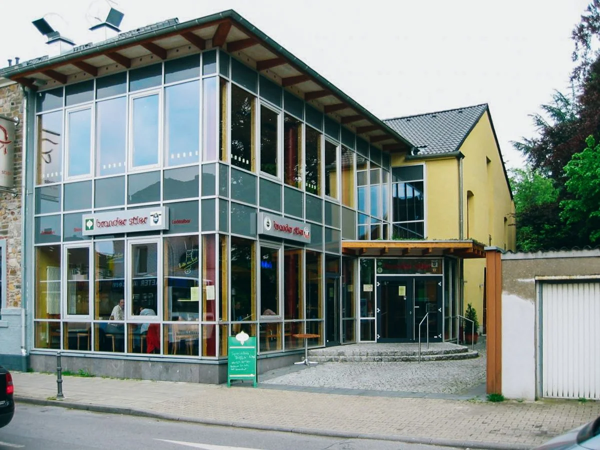 Ein modernes Gebäude mit großen Glasfenstern und gelber Wand, vermutlich ein Restaurant oder Café, mit einem kleinen Außenbereich und einem grünen Piktogramm auf einem Schild vor dem Eingang.