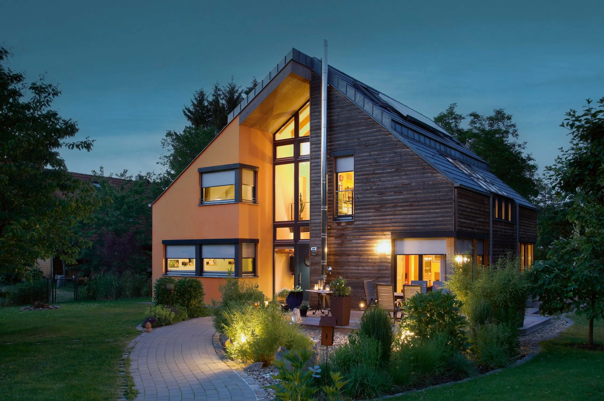 Moderne zweistöckige Holzhybridvilla mit großen Fenstern und Terrasse bei Nacht, umgeben von Grünflächen und Lichter im Garten