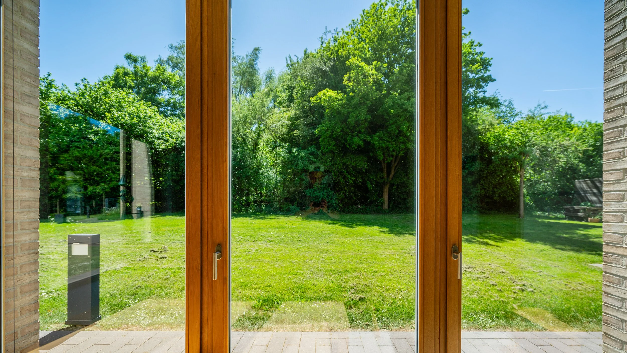 Blick durch eine große Glastür auf einen grünen Garten mit Bäumen, Rasen, einem kleinen Weg und einer Hecke bei sonnigem Wetter.