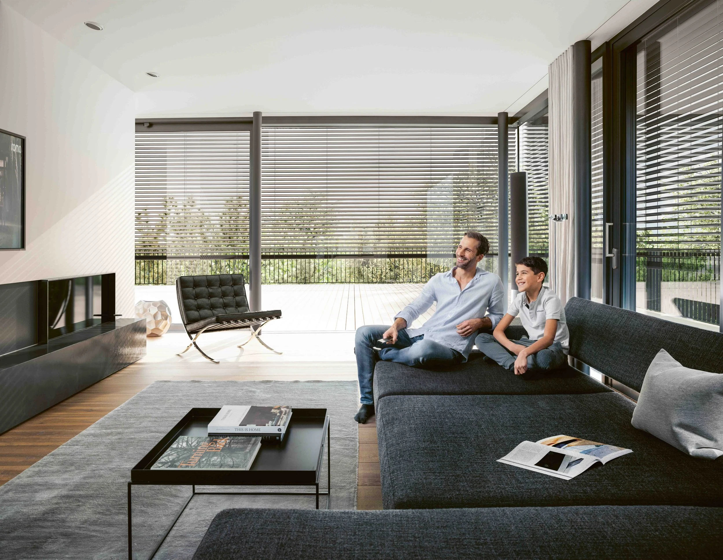 Ein modern eingerichtetes Wohnzimmer mit großen Fenstern, einem schwarzen Sofa, einem Kind und einem Erwachsenen, die gemeinsam sitzen, und einem Couchtisch mit Büchern, bei Tageslicht.