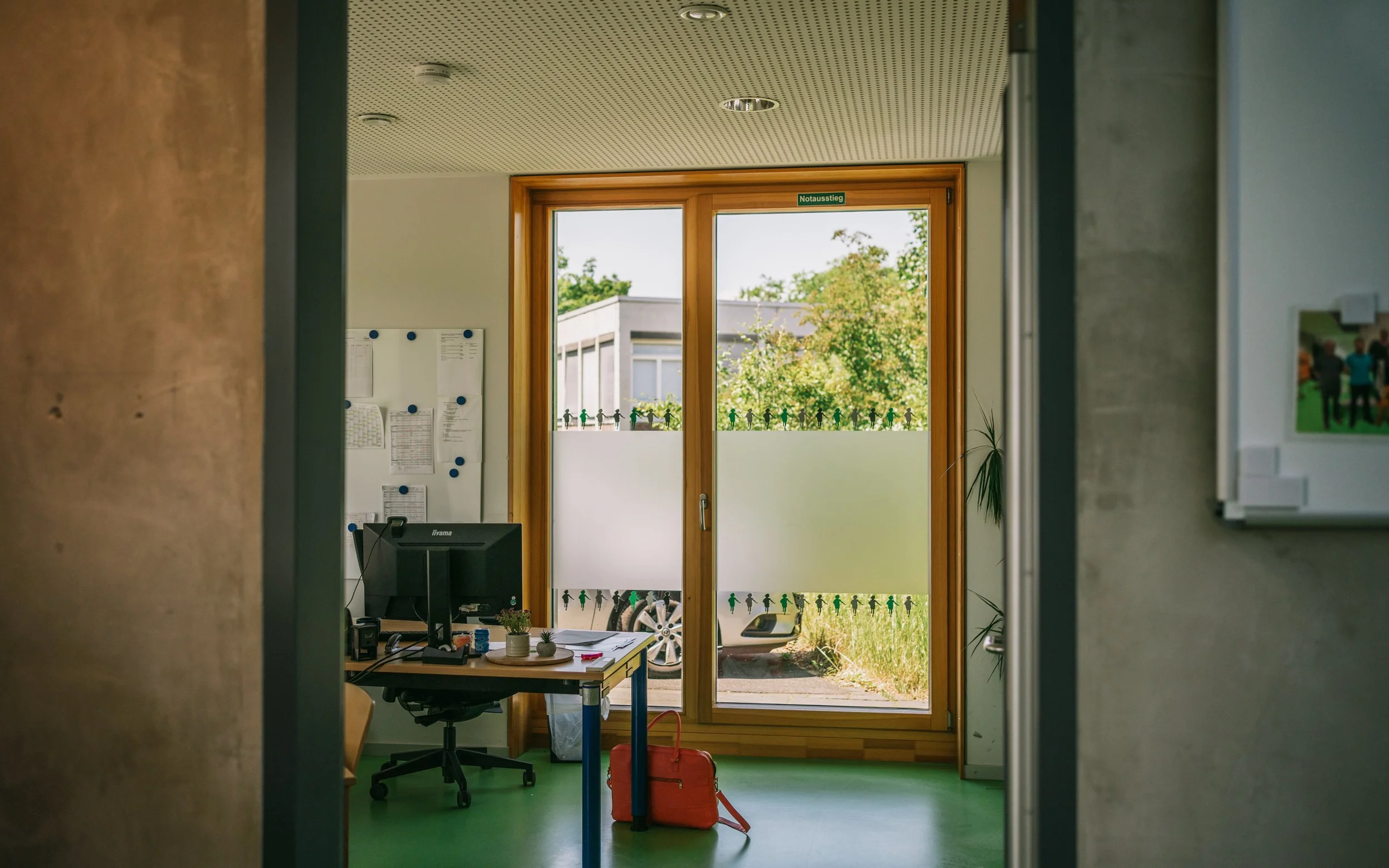 Büro mit Blick nach draußen durch große Glastür, Schreibtisch mit Computer, Pinnwand mit Dokumenten, Pflanzen und ein roter Aktenkoffer auf dem Boden.