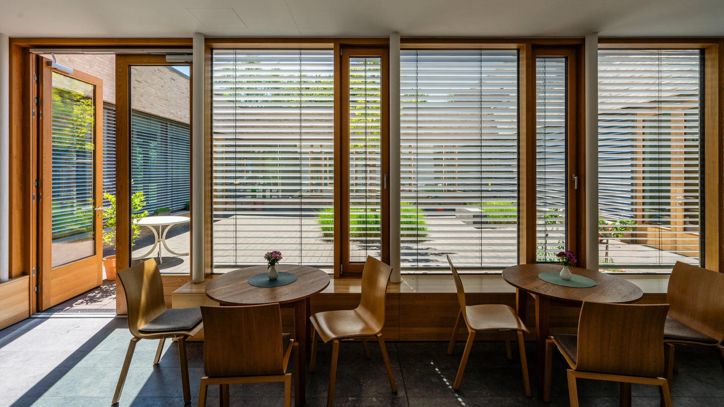 Innenraum mit Holztischen und Stühlen, große Fenster mit Jalousien, Blick nach draußen auf einen Garten mit Pflanzen, offener Ausgang auf eine Terrasse.