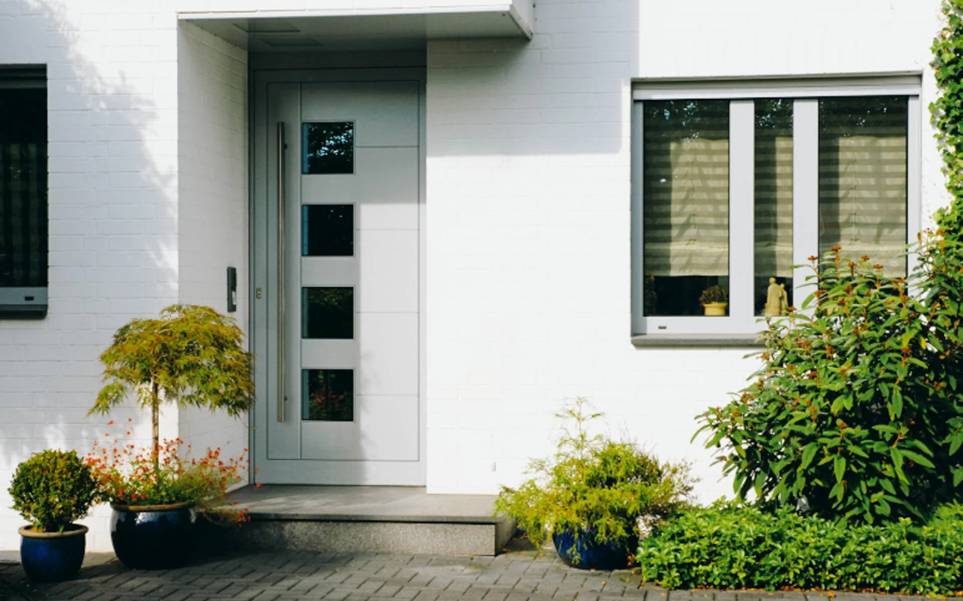 Weiße Hausfront mit Eingangstür und Fenster, umgeben von grünen Pflanzen und topfblumen, sonniger Tag.