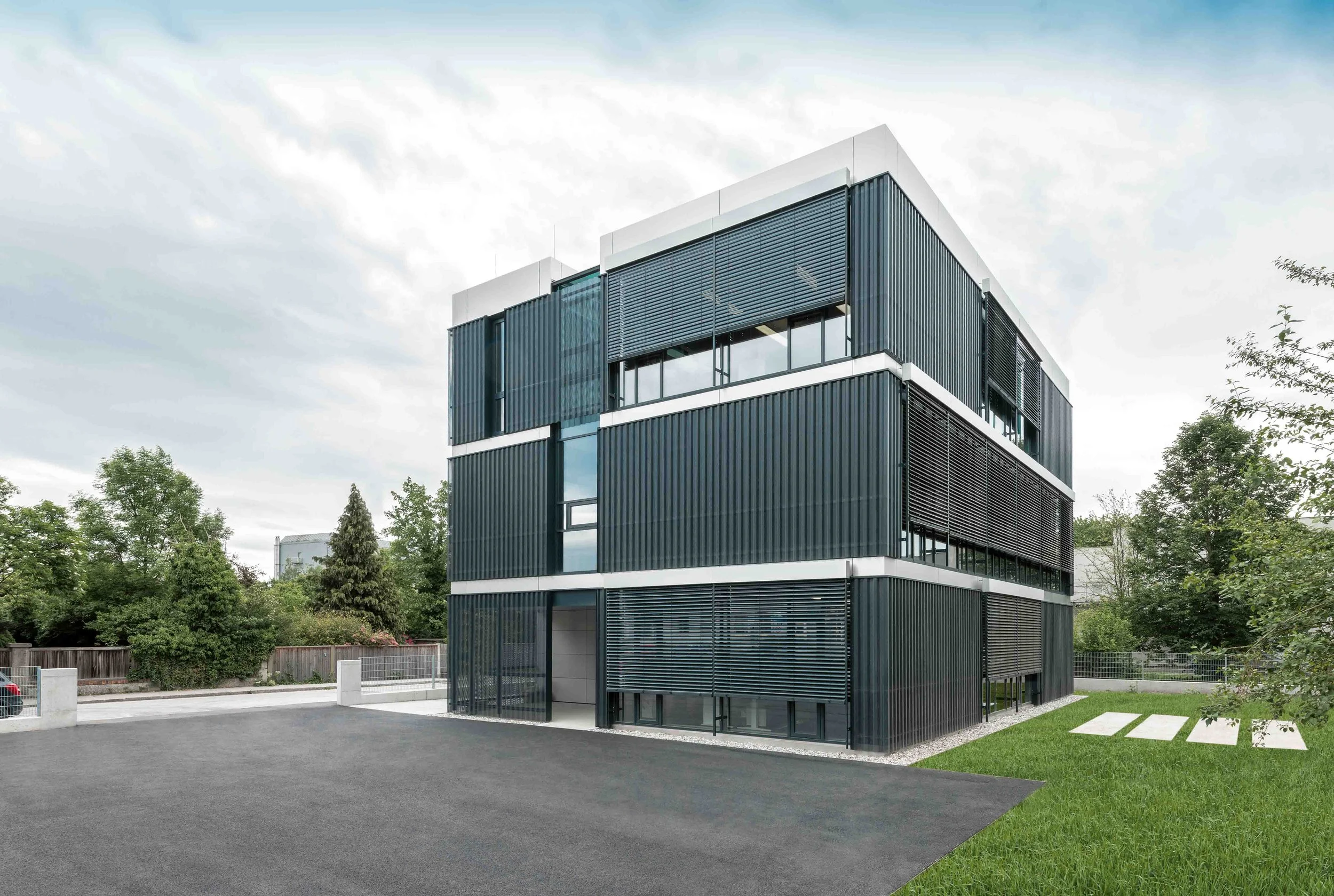 Modernes mehrstöckiges Bürogebäude mit dunkler Fassade und großen Fenstern, umgeben von Bäumen und einem gepflegten Rasen, bei bewölktem Himmel.