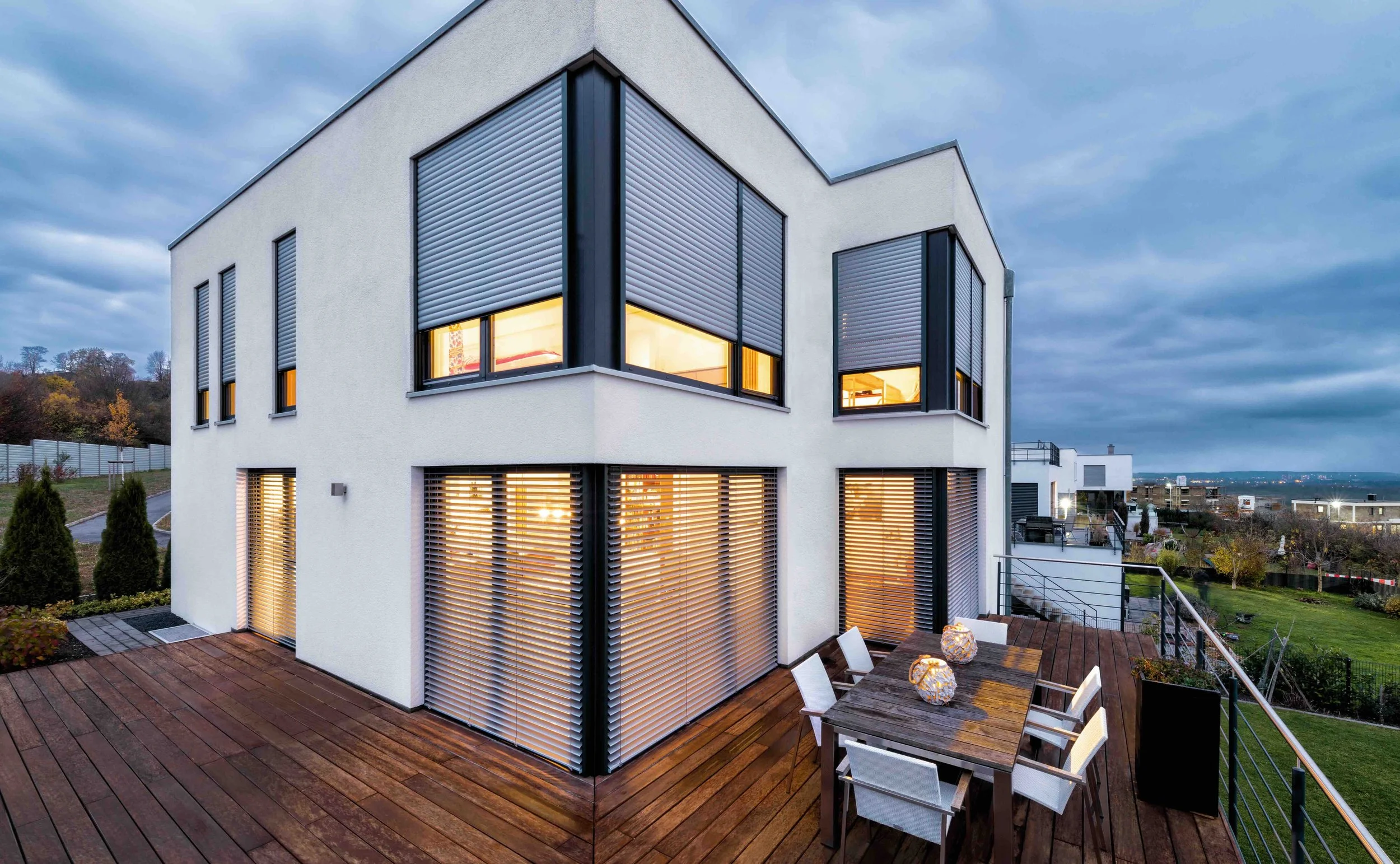 Moderne weiße Doppelhaushälfte mit großen Fenstern und Rollläden, Holzdachterrasse mit Esstisch und Stühlen bei bewölktem Himmel.