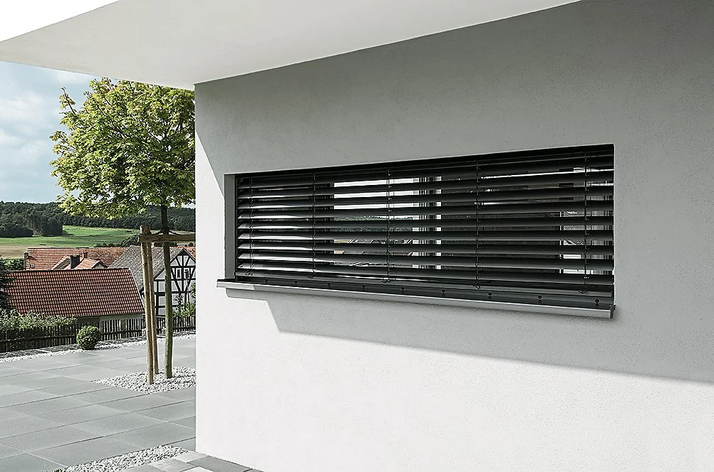 Moderne weiße Hausfassade mit horizontalen schwarzen Dachfensterläden, im Hintergrund Garten mit Baum und Blick auf ländliche Umgebung.