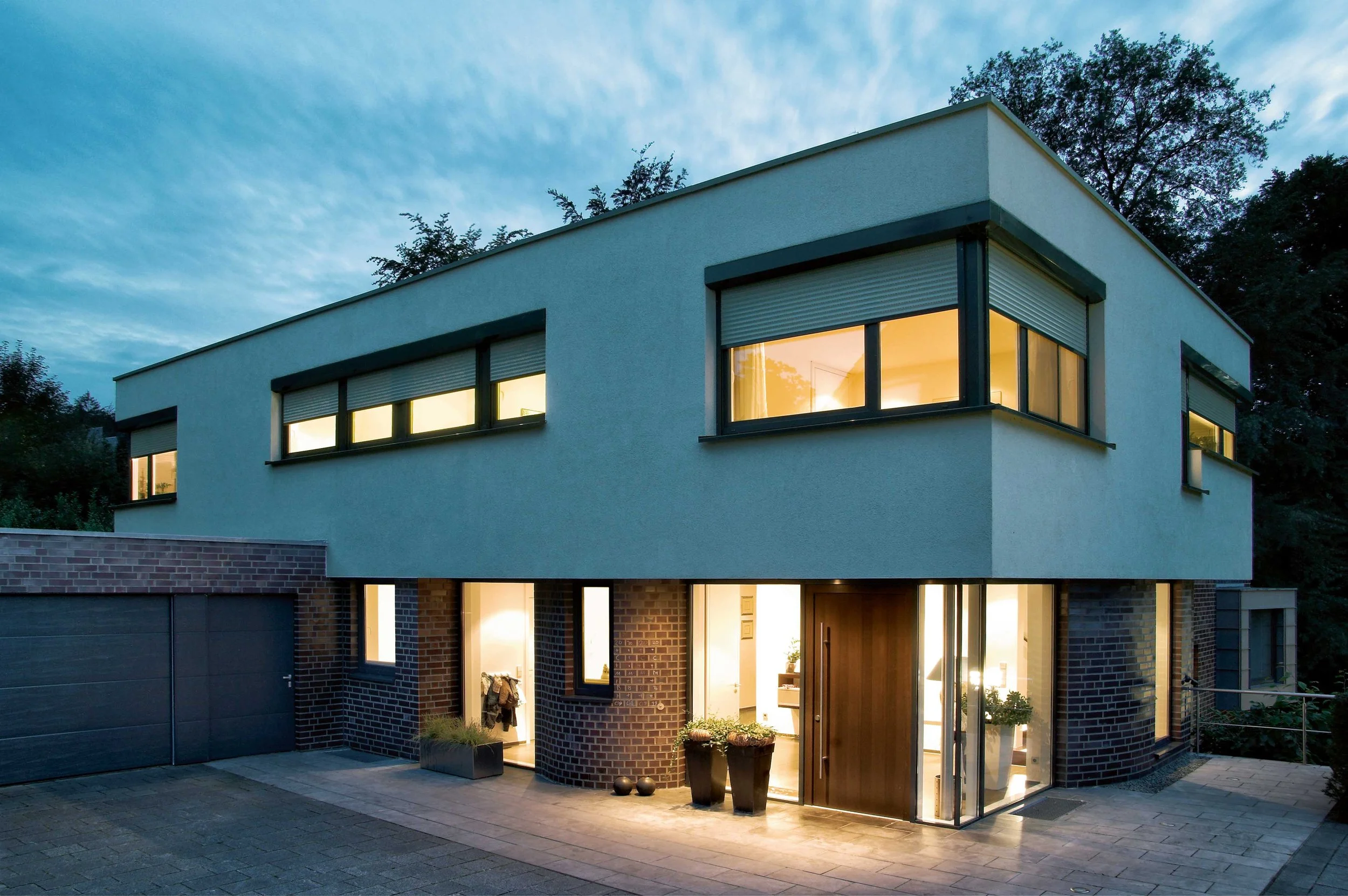 Modernes zweistöckiges Haus bei Dämmerung mit beleuchteten Fenstern und Eingangstür, umgeben von Bäumen.