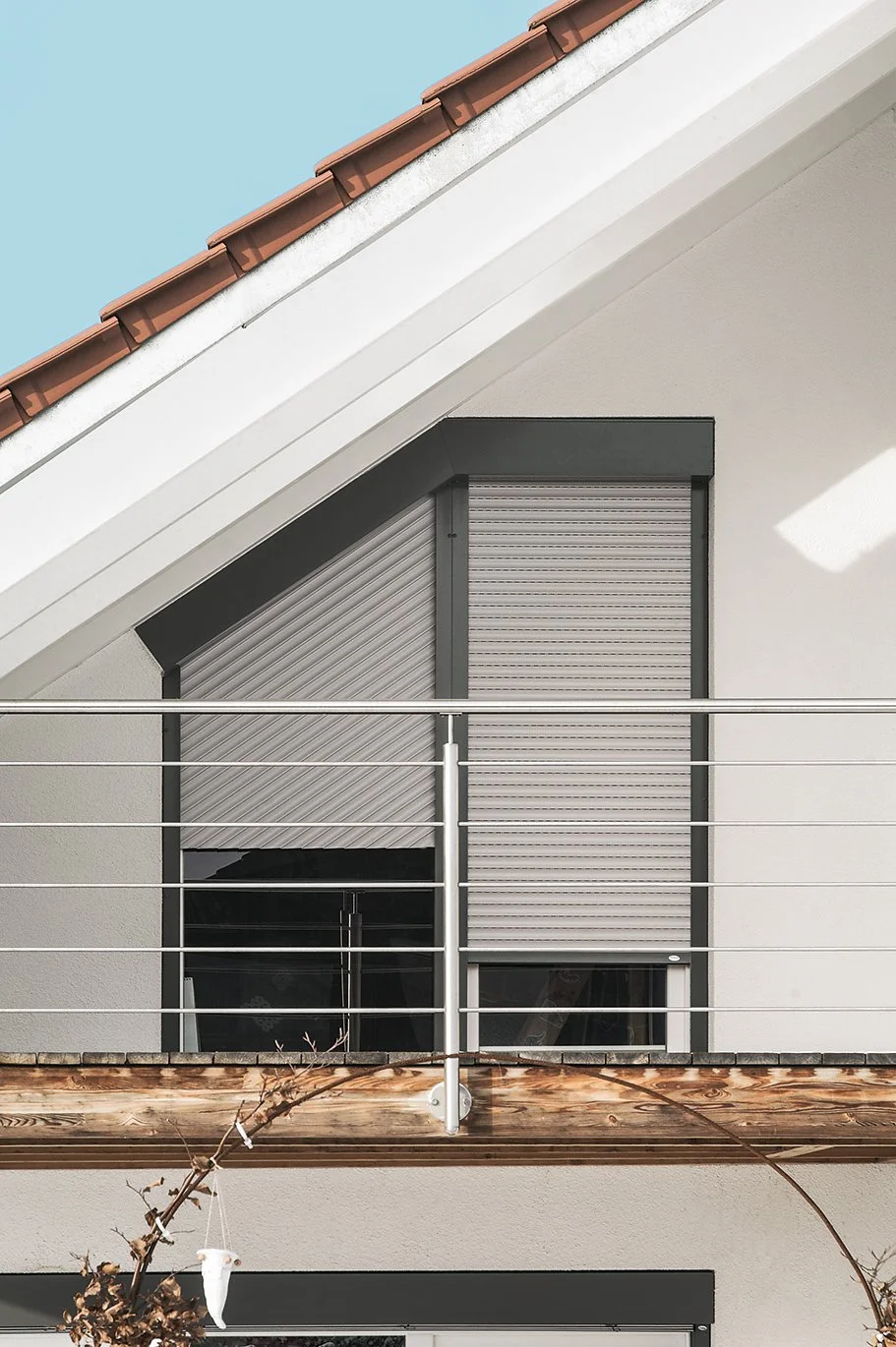 Ansicht eines modernen Hauses mit Balkon, rollbare Fensterläden und rotbraune Dachziegel unter blauem Himmel.