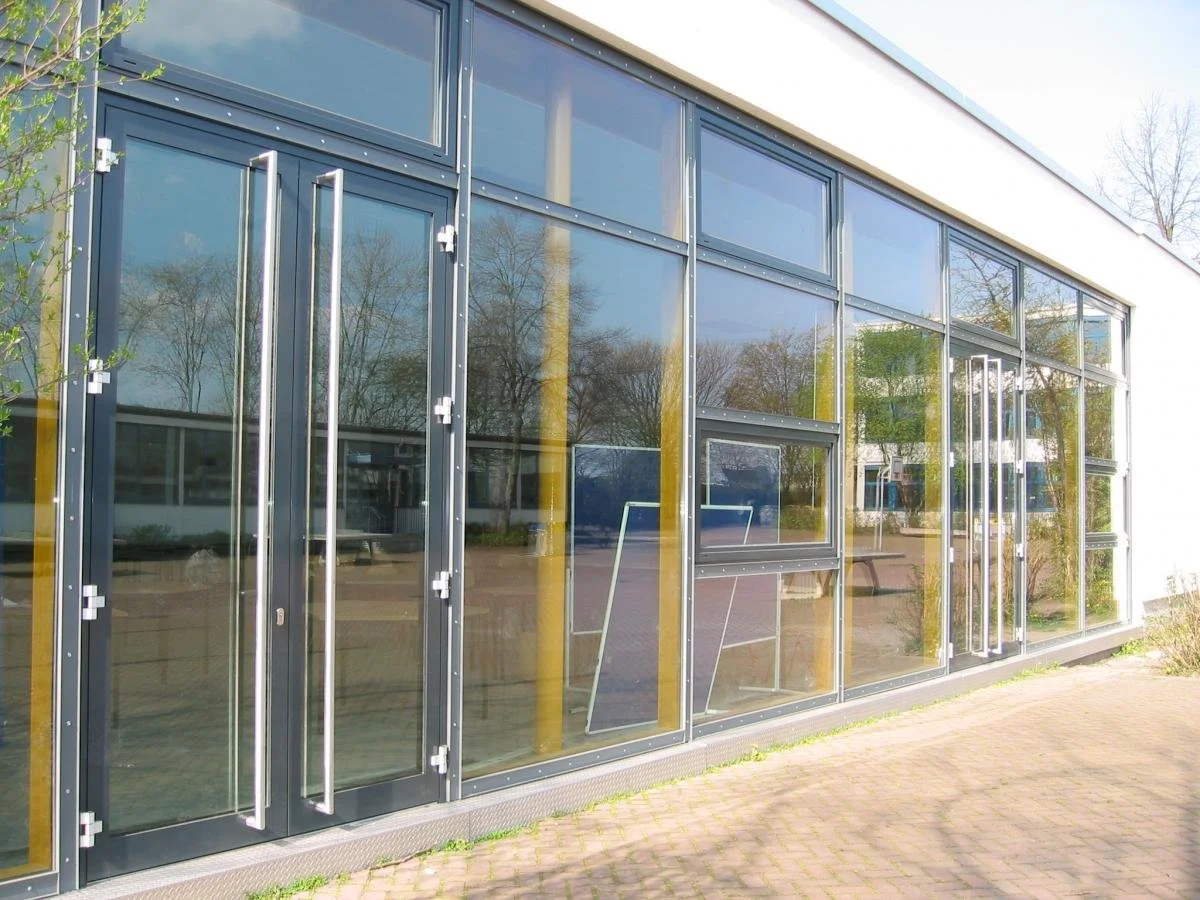 Moderne Glasfassade eines Gebäudes mit Passagiertüren und Fensterelementen, reflexionen von Bäumen und Himmel im Glas.