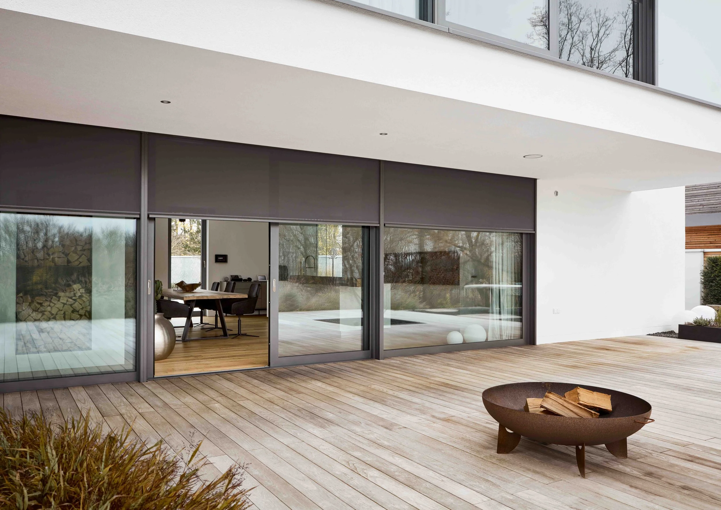 Moderne Terrasse mit Holzdecke, große Fenster und Holzboden, Blick ins Innere mit Esstisch, und im Vordergrund ein Feuerkorb mit Holz. Außenbereich mit Pflanzen und weißen Dekorelementen.