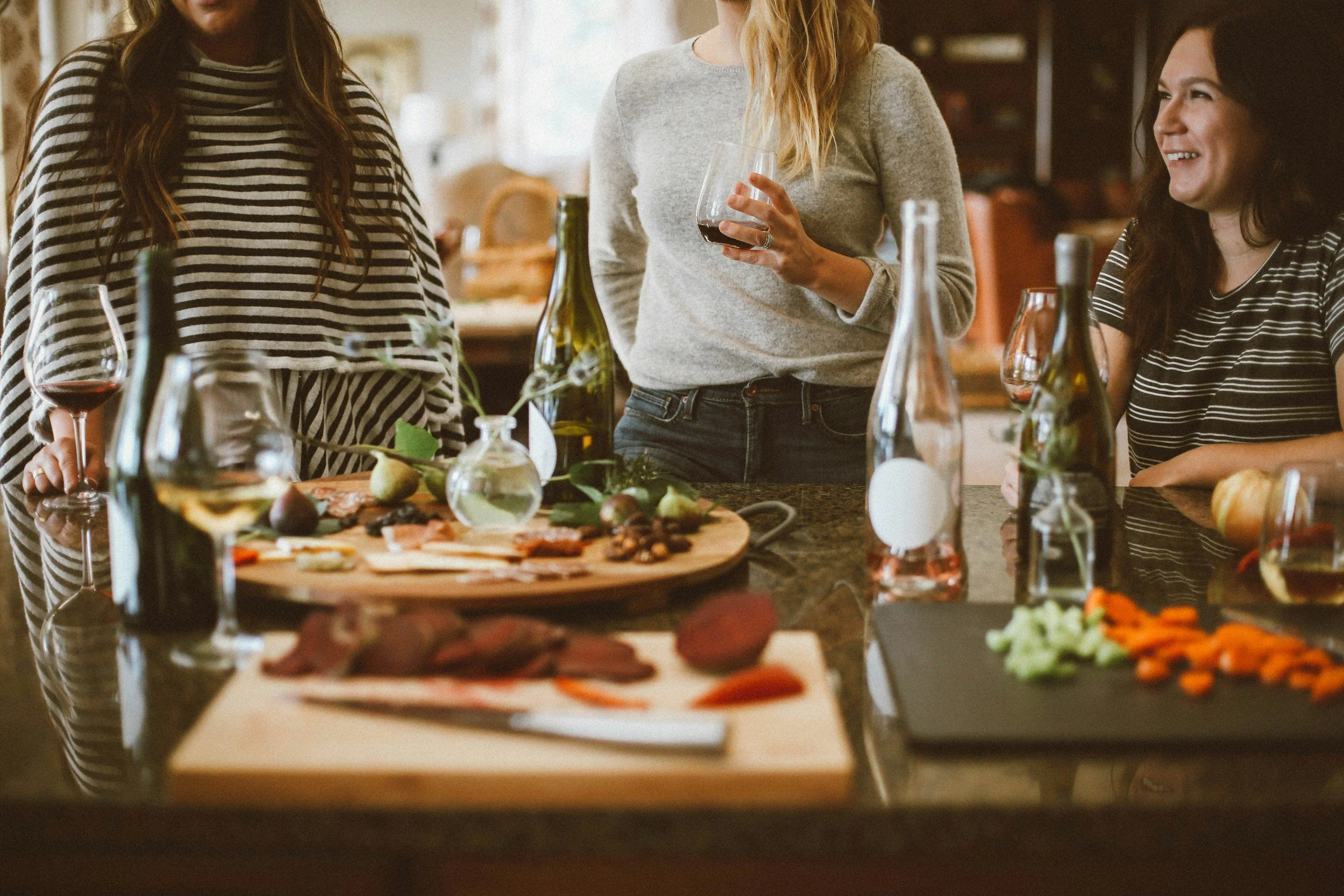 Three women gathered around a kitchen island enjoying wine and charcuterie, with food, bottles, and glasses on the counter.