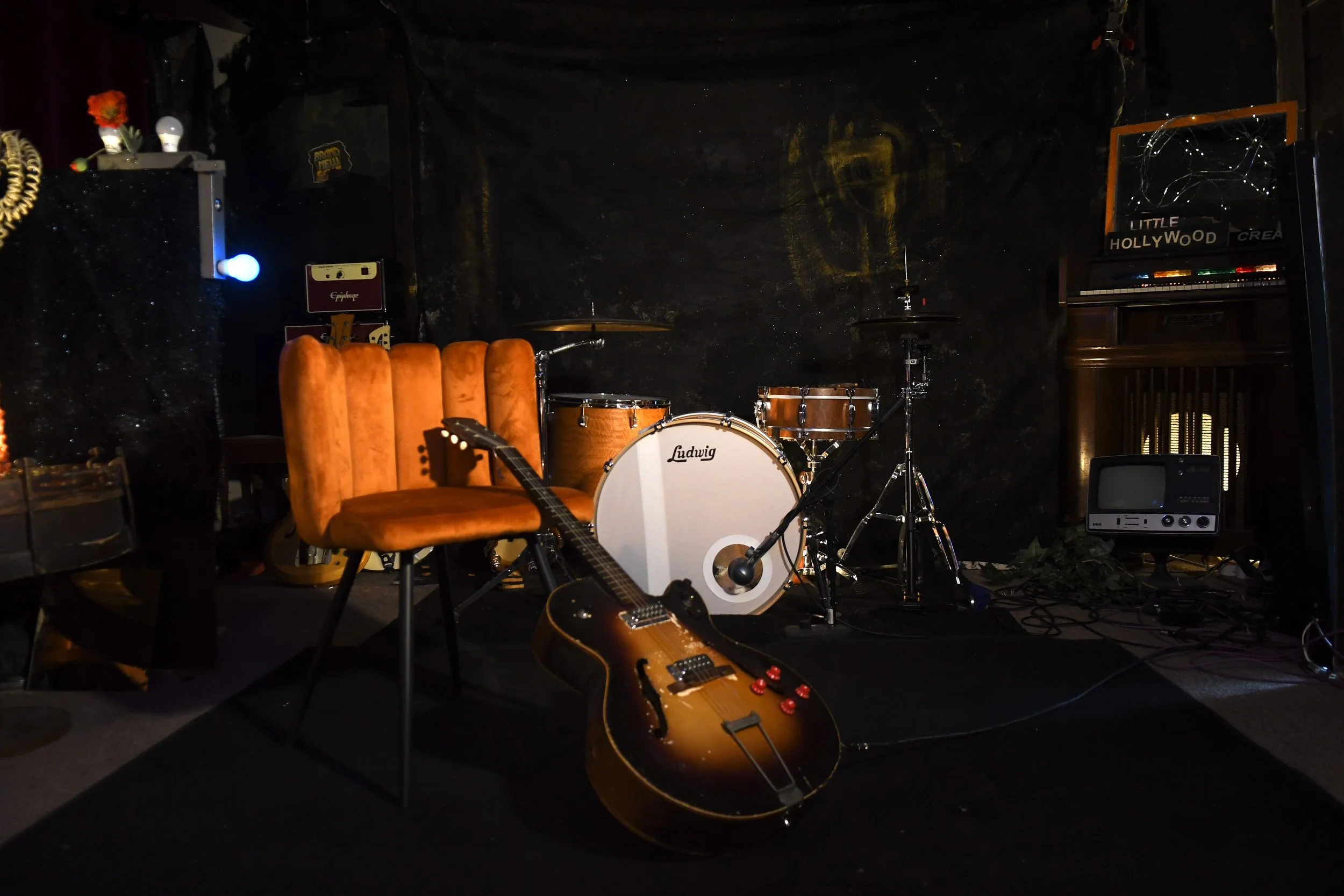 Music stage setup with drums, guitars, and an amplifier in a dark room with black walls and minimal lighting.