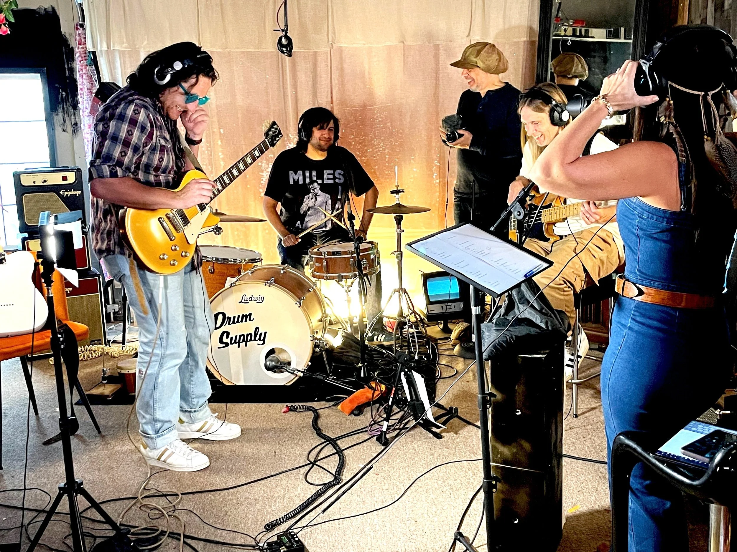 Group of musicians recording in a studio, playing guitars and drums, with professional microphones and headphones, and a music stand with sheet music.