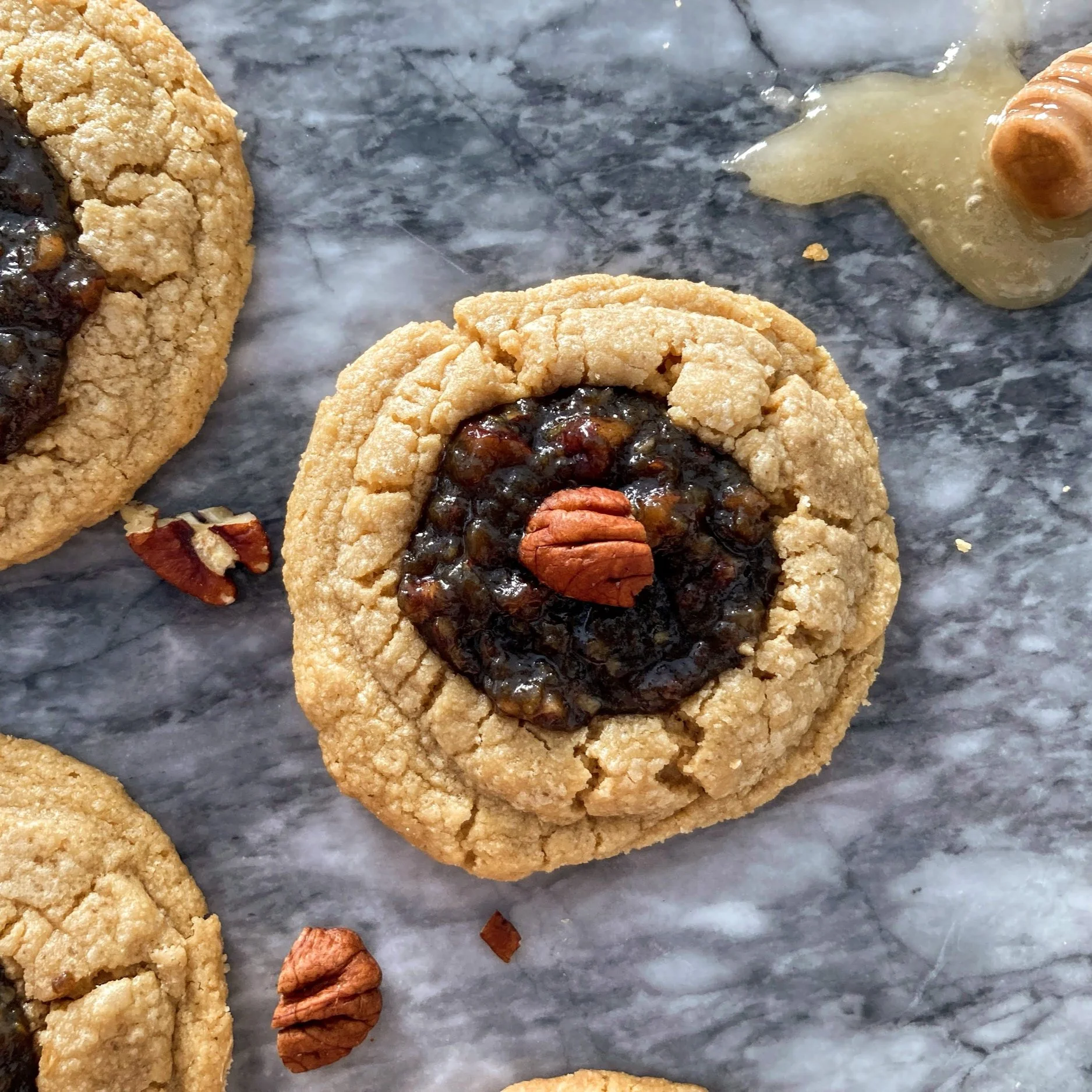 Close-up of a cookie with a pecan on top and pecan pie filling inside, placed on a marble surface with some nuts and honey nearby.