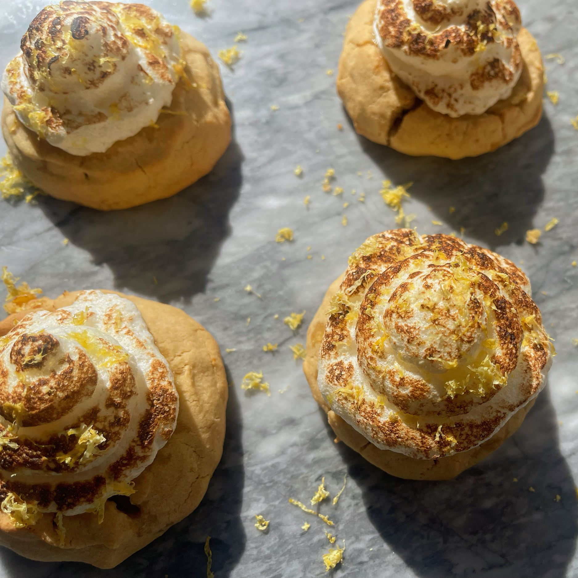 Four lemon-flavored cookies with toasted meringue on top, placed on gray parchment paper with lemon zest scattered around.
