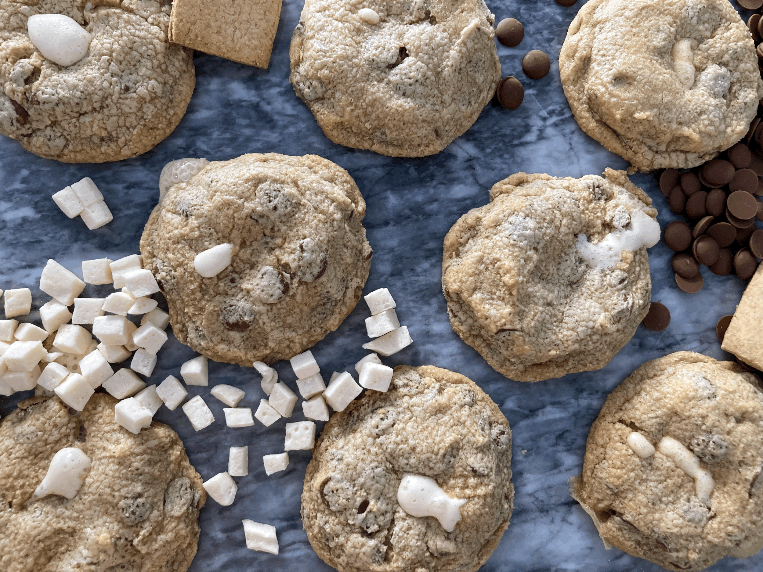 Several homemade gluten free smores cookies with chocolate chips and marshmallows on a marble surface.